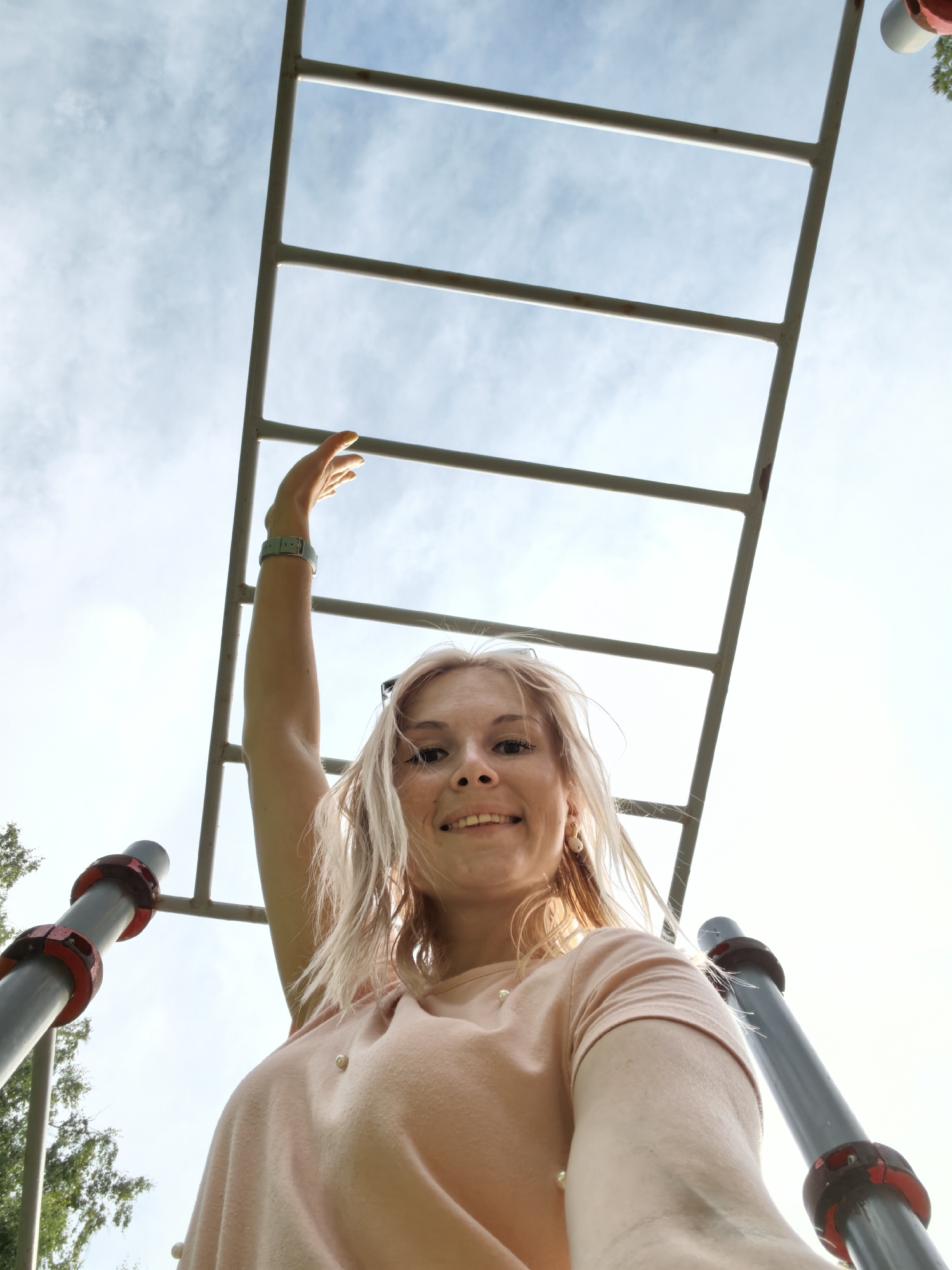 A low-angle, full-body shot shows a young, blonde woman smiling as she reaches up towards a monkey bar structure. She is wearing a light pink t-shirt adorned with small pearl-like embellishments and a blue digital watch on her left wrist. Her hair is slightly windswept, and she is wearing earrings.
The monkey bar structure is made of metal bars, which are a light gray color with some rust spots visible on the edges. The woman is positioned in the foreground, looking up at the camera. The background consists of a bright, cloudy sky with patches of blue.
On the left and right sides of the frame, parts of the metal support poles of the monkey bar are visible. These poles are dark gray with red accents. In the lower left corner, some green foliage and tree branches are visible, suggesting an outdoor setting, likely a park or playground.
The sunlight appears to be bright, indicating it is daytime, possibly late morning or early afternoon. The overall mood of the image is cheerful and active, with the woman appearing energetic and happy. The specific location is Sterlitamak, Russia. There is no visible text in the image.