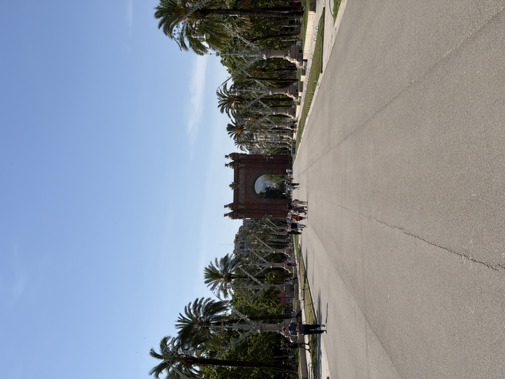 The image depicts a view looking down a wide, paved pathway lined with palm trees and ornate lampposts. In the distance, an ornate brick archway stands prominently. Several people are visible walking along the pathway, some in small groups and others individually. The sky is clear and blue. This scene is characteristic of the Arc de Triomf in Barcelona, Spain, a triumphal arch located at the northern end of the Passeig de Lluís Companys.