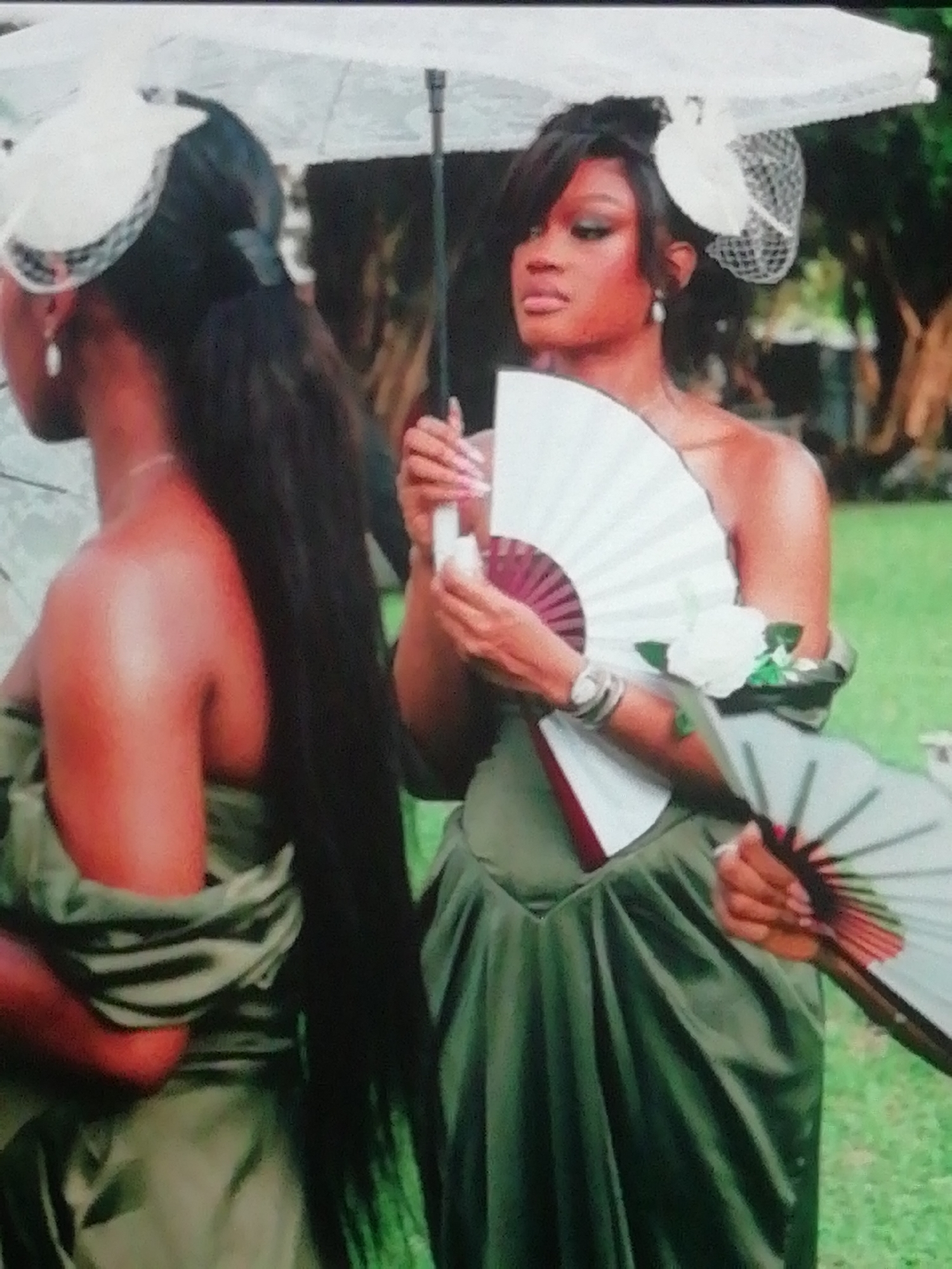 Two Black women are depicted outdoors, dressed in matching olive green strapless gowns, suggesting they are attending a formal event, possibly a wedding or garden party. The scene is set against a lush green, natural background, indicative of a tropical or sub-tropical climate like Enugu, Nigeria.
One woman is shown from the back, her long, dark hair styled in a ponytail. She wears a white fascinator or hat on her head and a pearl drop earring.
The second woman, in the foreground, is positioned slightly to the left, looking away from the camera with a seemingly serious or contemplative expression. Her dark hair is styled in an updo with bangs, and she wears a white fascinator adorned with netting. She holds a white and maroon paper fan in her right hand, with another fan partially visible in her left hand, suggesting warm weather. Her long, light pink manicured nails are visible as she holds a small white object, possibly a spray bottle. Her left wrist is adorned with a watch and multiple bracelets, and a white rose corsage is pinned to her left shoulder.
A large white parasol provides shade over both women, further hinting at a sunny, daytime event. The overall atmosphere suggests an elegant outdoor gathering where guests are dressed formally and prepared for the heat. No visible text is present in the image.