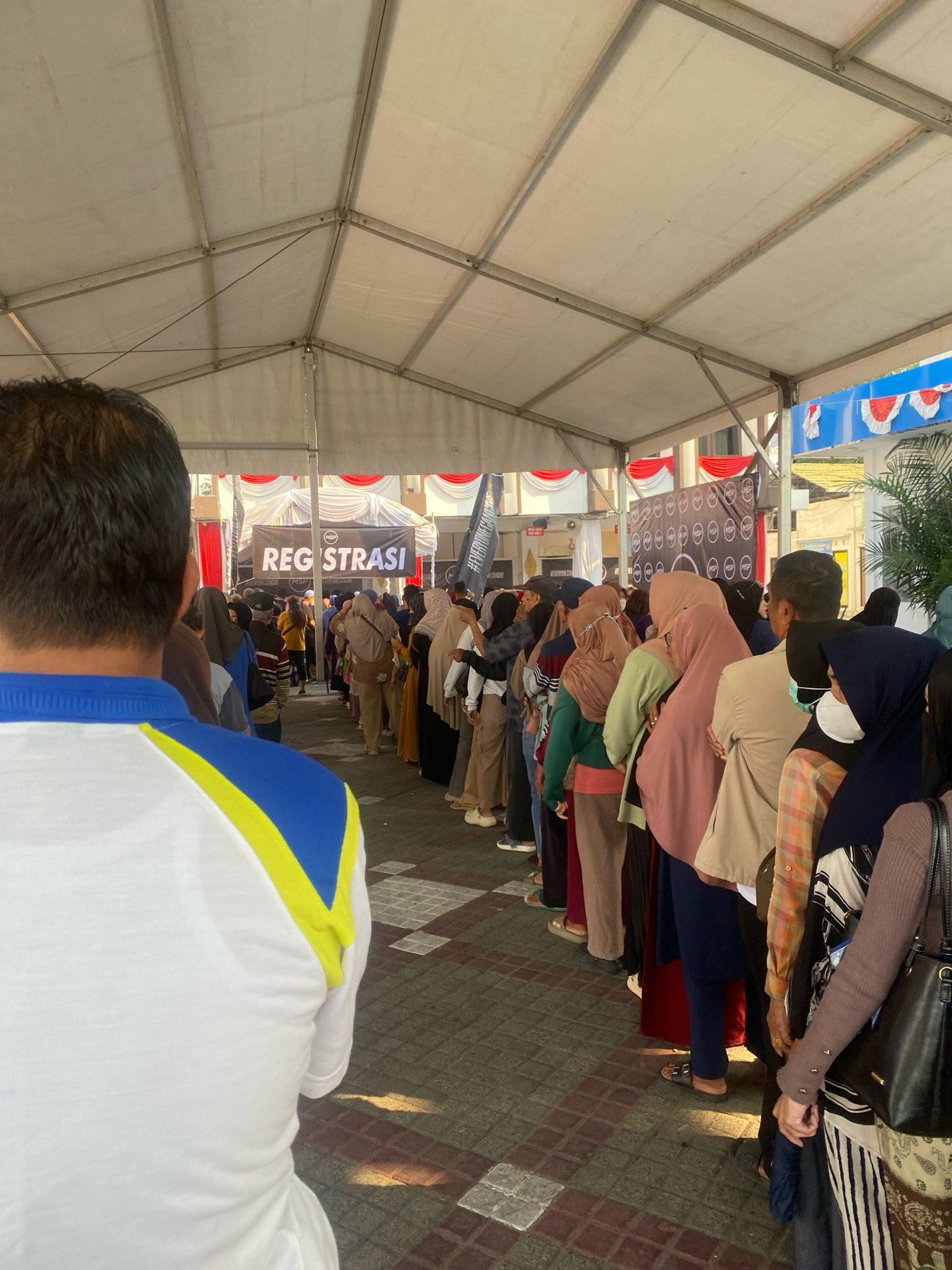 The image shows a queue of people, predominantly women wearing hijabs, waiting in a line under a large tent. A banner in the background reads "REGISTRASI". The ground is paved with interlocking bricks. In the foreground, the back of a person wearing a white shirt with blue and yellow accents is visible. The event appears to be an outdoor registration or check-in process. The context provided states this is in Banjarmasin, Indonesia.