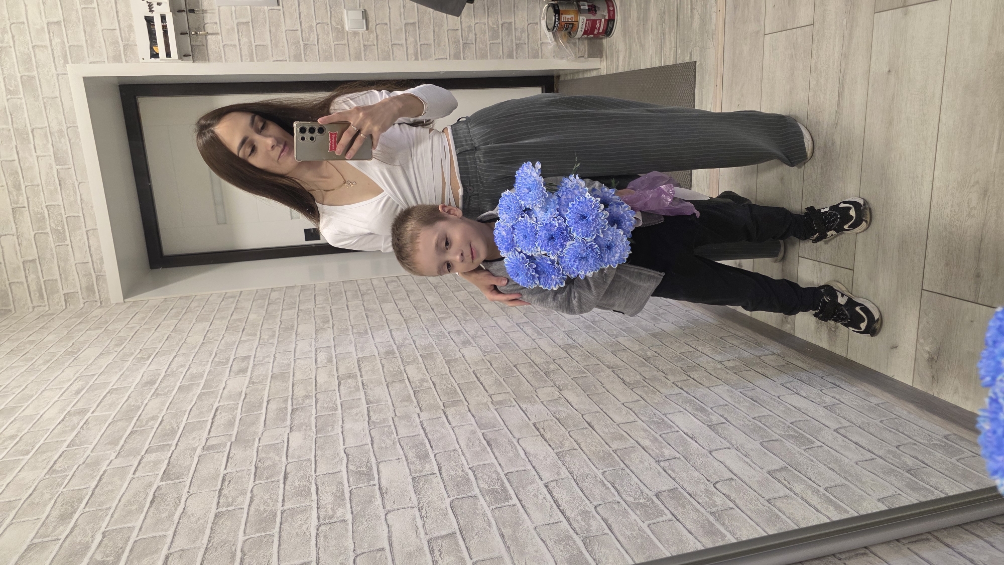 In an indoor setting, a woman with long brown hair stands in front of a brick-patterned wall, taking a mirror selfie. She is wearing a white wrap top and high-waisted pinstripe pants. To her left, a young boy stands holding a bouquet of blue flowers. He is wearing a grey jacket, black pants, and black and white sneakers. The woman has her arm around the boy's shoulders. The environment appears to be a hallway or entryway, with a door visible in the background and a mirror on the left side of the frame.