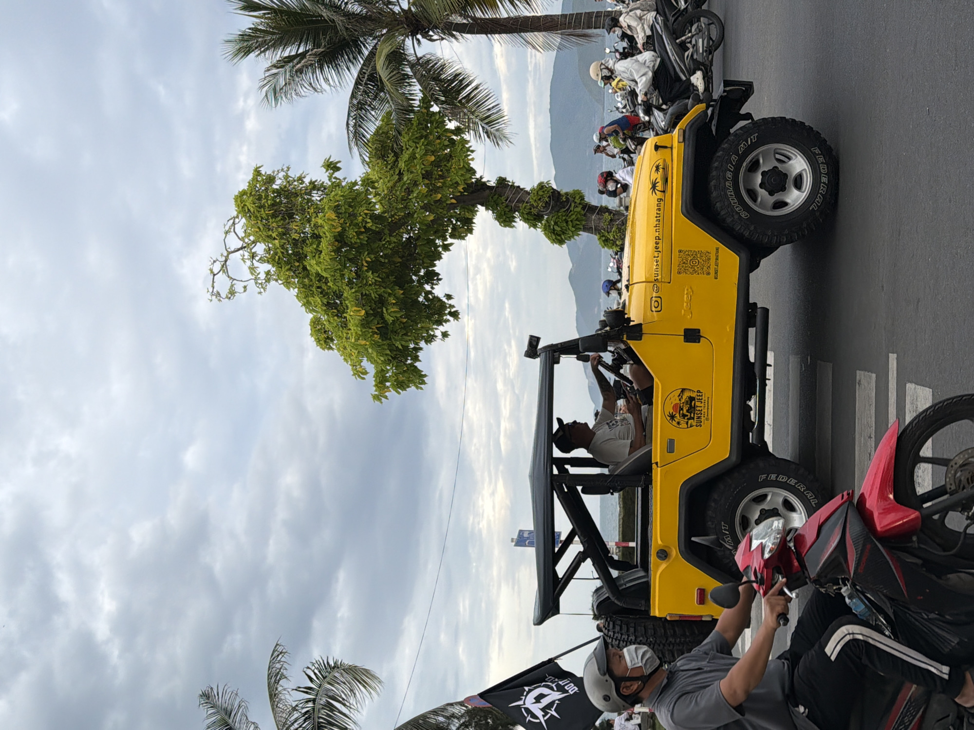 In Nha Trang, Vietnam, on a cloudy day, a yellow Jeep is parked on a street. A man wearing a grey shirt, black pants, and a face mask is riding a red and black motorcycle in front of the Jeep. The Jeep has a black roof and roll cage, and a spare tire is mounted on the back. A black flag with a white logo is planted to the left of the Jeep. Several people are visible on motorcycles behind the Jeep, some wearing helmets and masks. Palm trees are on the left and right sides of the frame, and in the background, mountains and the sea are visible. The Jeep has logos on its side, including "SUNSET JEEP" and a QR code.