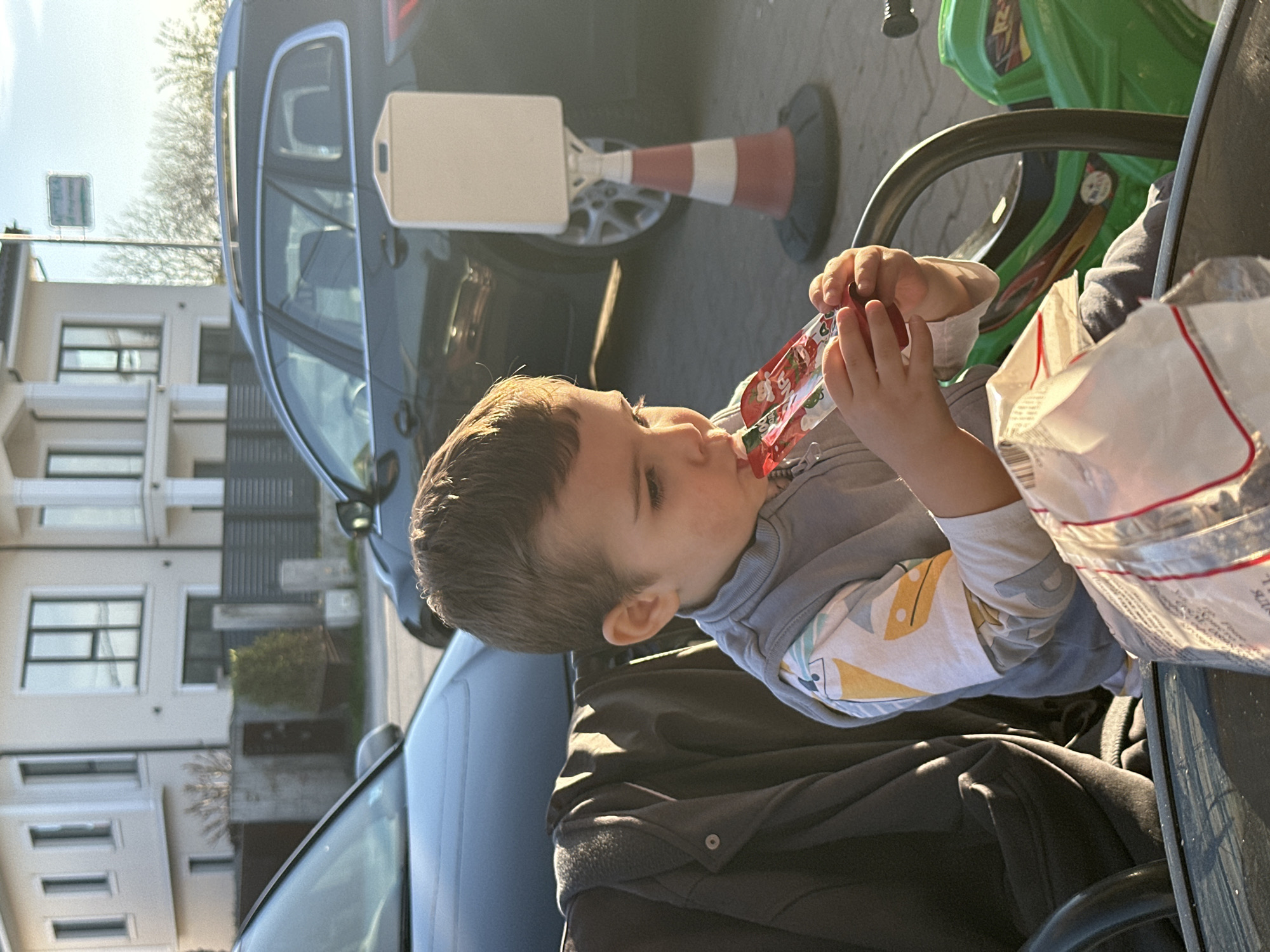 A young child is drinking from a pouch while seated in a location with outdoor seating, possibly a cafe or restaurant. A dark-colored car is parked behind the child, and a traffic cone with a white sign is positioned near the car. Buildings are visible in the background, including a modern-looking residence with multiple windows. Another car is to the left of the child, parked on a street. The image does not contain sufficient information to determine whether the scene is in Plovdiv, Bulgaria.
