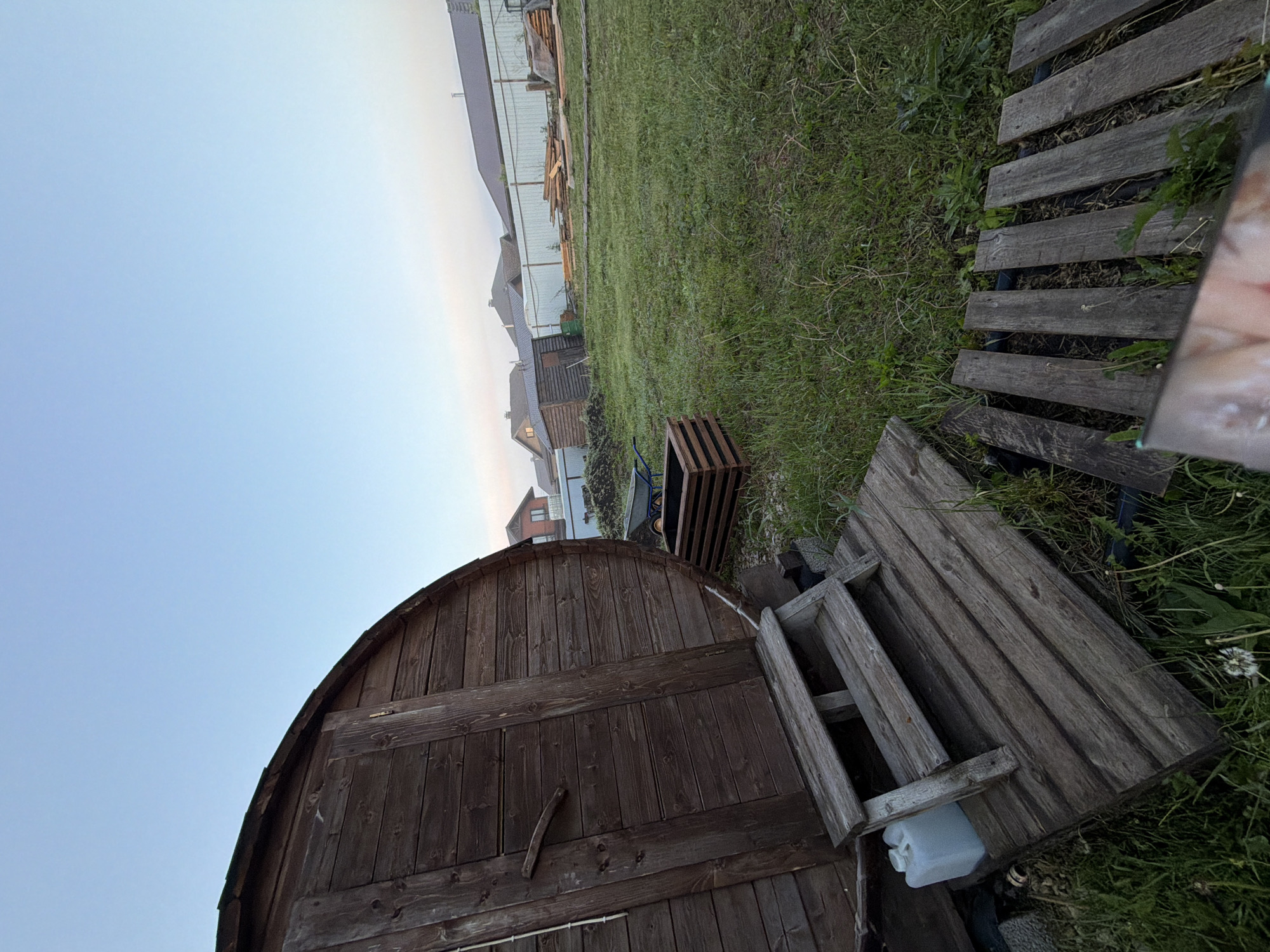 The image, originally oriented in portrait mode and rotated 90 degrees counter-clockwise, depicts a rural or suburban backyard scene in Большая Шильна, Russia. The sky, visible across the top and left, is clear and light blue, indicating daytime, likely early morning or late afternoon due to the soft, diffused light and absence of harsh shadows.
In the foreground, a dark brown, barrel-shaped wooden structure, possibly a shed or a small outbuilding, occupies the lower-left portion of the frame. Its rounded roof and vertical wooden planks are clearly visible. Next to it, on the right, a wooden panel or gate lies flat on the ground, and beneath it, a white plastic container is partially visible. Further to the right, a makeshift pathway of weathered wooden planks extends into the grassy area. A faint reflection, possibly of a person's face or hand from a smartphone screen, is visible on the far right edge of the image.
The midground features a patch of unkempt green grass with some weeds. Towards the center-right, a wooden slatted crate or planter box sits on the ground, with a blue object (possibly a wheelbarrow handle) partially seen behind it. In the background, a light-colored metal fence separates the yard from a residential area. Beyond the fence, several houses with dark roofs are visible, and a pile of stacked wooden planks or building materials can be seen near the fence.
No specific activity is taking place; the scene is static. There are no clearly visible individuals, apart from the possible reflection. No discernible text is present in the image.