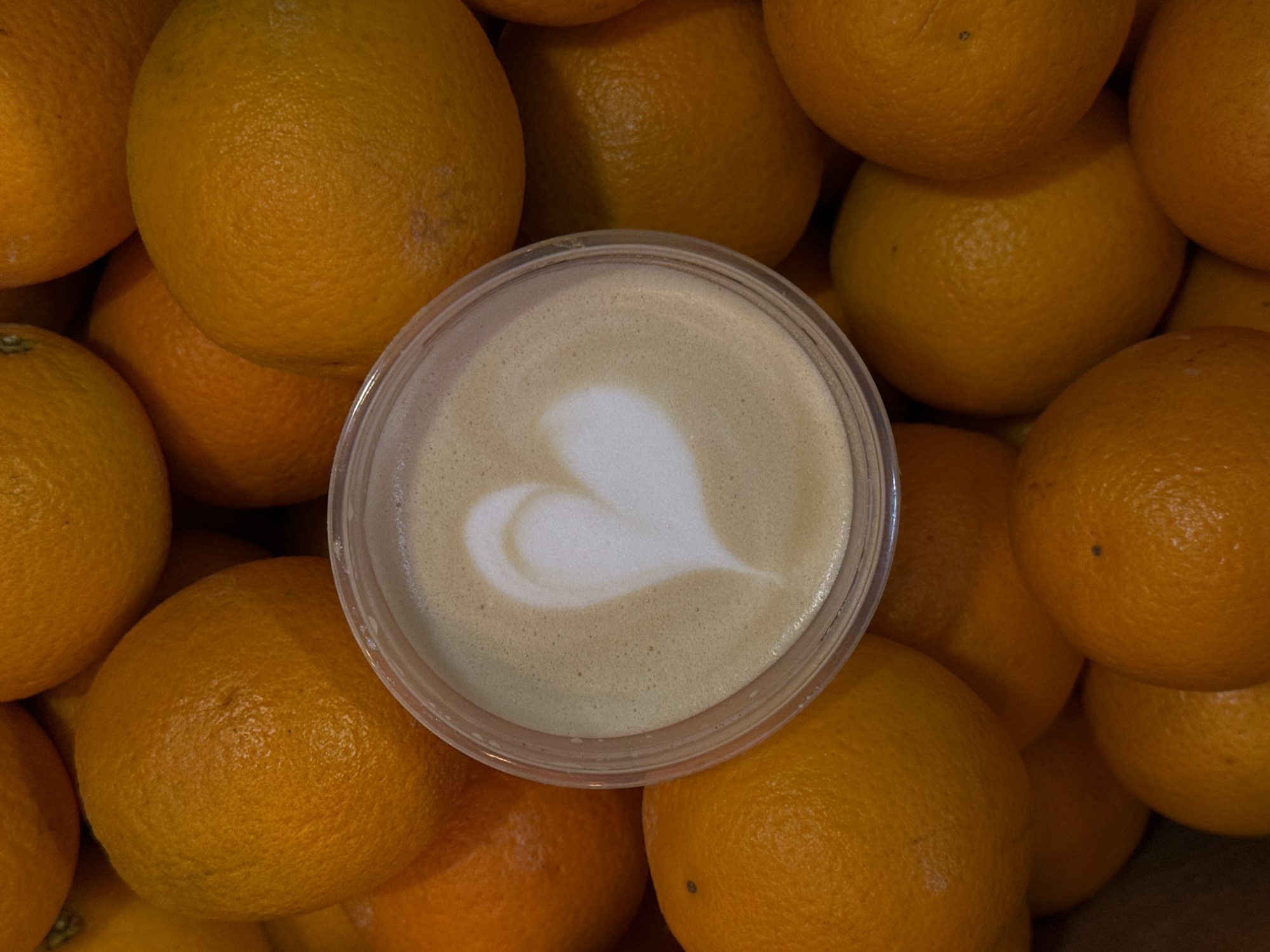The image shows a clear plastic container filled with coffee with latte art in the shape of a heart, placed in the center of a pile of oranges. The oranges are a vibrant orange color and appear fresh. The coffee is a light brown color with a creamy foam topping. The background is blurred, but it appears to be a market or store setting, given the abundance of fruit. There are no people or animals visible in the image. The location in the city of Moscow, Russia is not specified.