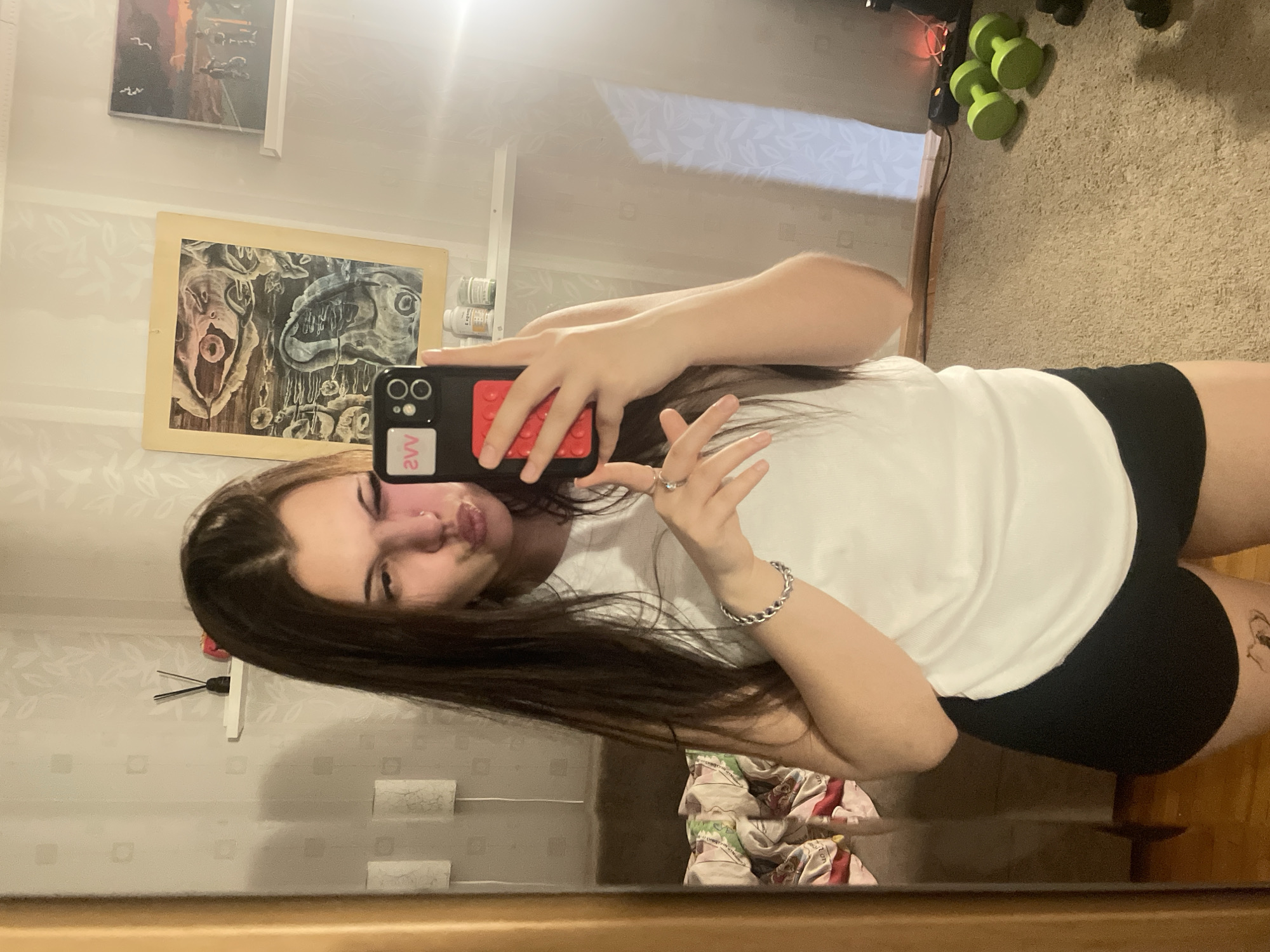 The image displays a mirror selfie taken by a young woman in Saint Petersburg, Russia. She has long, dark hair, wears a white t-shirt and black shorts or underwear, and is making a pouty expression. She holds a black smartphone with a distinctive red Lego-style back panel and a pink "2vv" sticker. On her left hand, she wears a ring, and a silver chain bracelet adorns her left wrist. A subtle tattoo is partially visible on her right thigh.
The reflection in the mirror reveals elements of the room's interior. Two framed artworks hang on the wall: an abstract landscape depicting a sunset or sunrise with birds, positioned above a more striking, darker piece featuring surreal, grotesque organic forms, including one that resembles an eye. A white shelf below this artwork holds several white cylindrical containers, possibly bottles of medicine or supplements. The floor is carpeted in a light brown, with a pair of green dumbbells and some black dumbbells visible. The walls feature light-colored wallpaper with a subtle patterned design. The lighting appears to be indoor, with some glare evident on the mirror's surface. The image is oriented sideways.