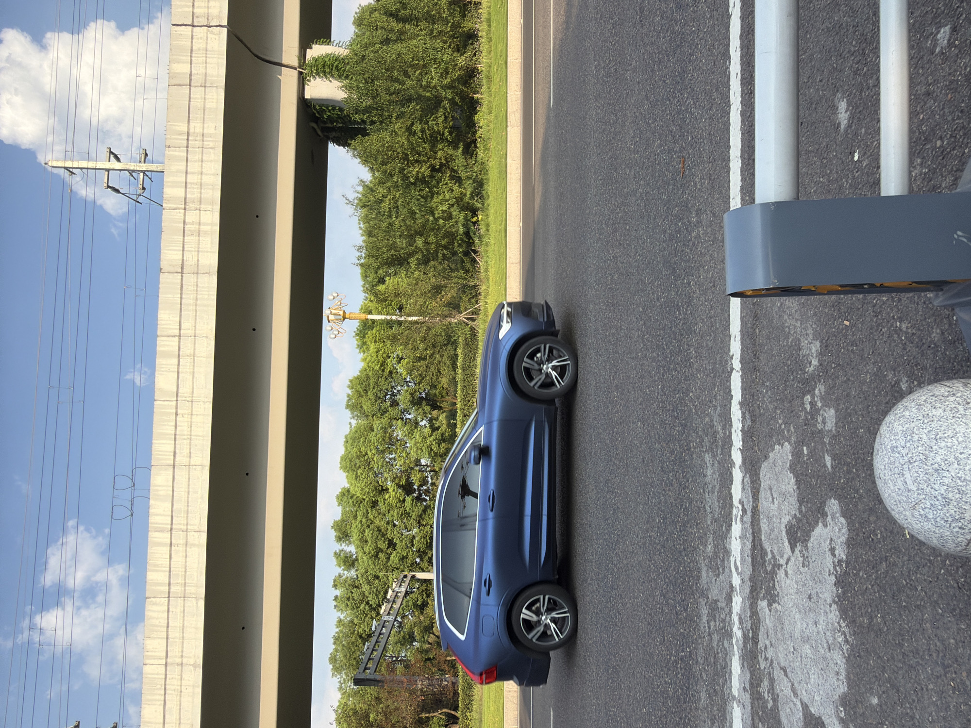 A blue Volvo XC60 is driving on a paved road. The car is in profile, moving from left to right. It has silver alloy wheels. The road has a white lane marking and some patches of wear and tear. In the background, there is a green hedge. Above the hedge, a concrete overpass supports overhead power lines. The sky is blue with scattered clouds. The scene appears to be in Jiangjin District, Chongqing, China, possibly near a major road or highway underpass.