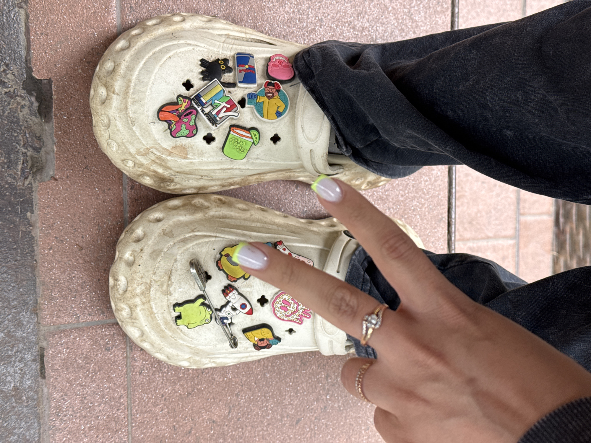 The image shows a close-up view of a person's feet wearing beige Crocs adorned with various colorful charms. The Crocs are placed on a tiled surface, with some stonework visible to the left. The person is wearing dark denim pants.
The charms on the left Croc include a green frog, a rocket ship, and a safety pin. The right Croc features a black cat, a Red Bull can, a pink skull, a person in a yellow jacket, a cocktail, and a colorful "MUSIC TELEVISION" charm. A hand with a manicure is visible in the lower right corner, making a peace sign and wearing two rings, one with a heart-shaped diamond.
The lighting suggests it is daytime, and the overall scene appears casual, possibly outdoors given the stonework. No specific time of day or weather conditions can be determined, and the location context of Naro-Fominsk, Russia, is provided but not visually evident in the image itself. The mood conveyed is playful and personalized due to the decorative charms on the footwear.