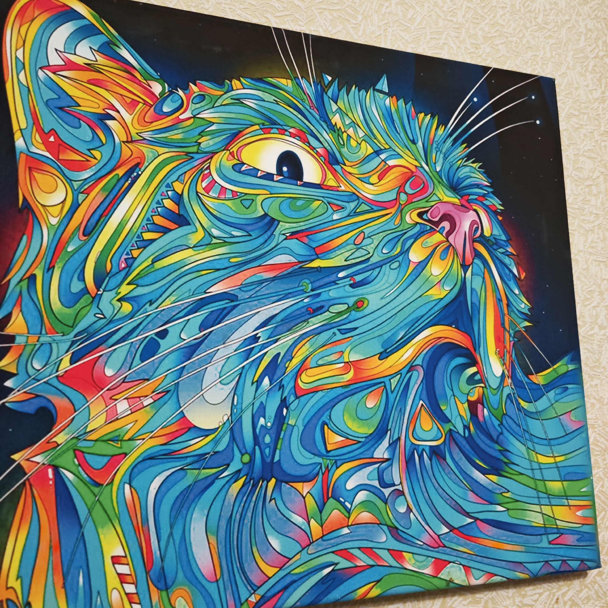 The image shows a colorful painting of a cat on canvas, seemingly mounted on a wall. The cat is depicted in a psychedelic style, with vibrant blues, yellows, greens, oranges, and reds forming swirling patterns and shapes to represent its fur, features, and whiskers. The background is dark, resembling a night sky with small dots possibly indicating stars. The wall behind the painting is a light beige color with a subtle texture. The painting appears to be viewed indoors. There is no indication of the location in Rostov, Russia.