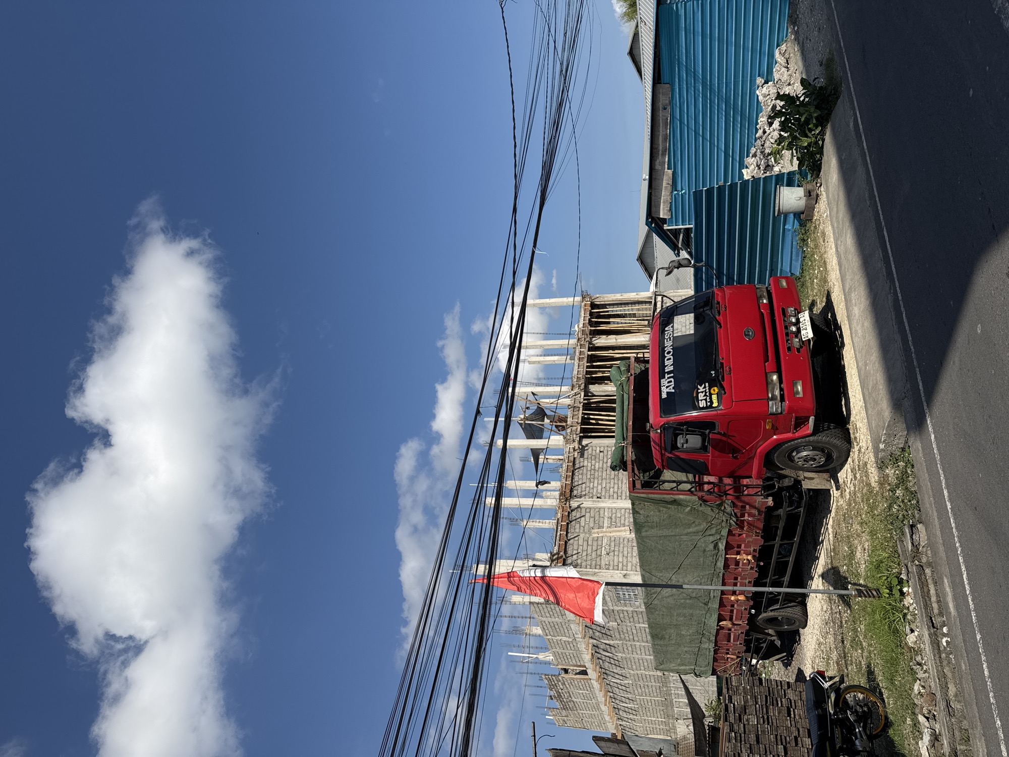 In the city of North Kuta, Indonesia, a red flatbed truck with a green tarp covering its cargo is parked on a dirt shoulder beside a paved road. A motorcycle is parked to the left of the truck. Behind the truck, a building under construction is visible, with concrete pillars and brick walls. An Indonesian flag, red and white, is prominently displayed on a pole in front of the construction site. Overhead, a tangle of electrical wires stretches across the bright blue sky, which is dotted with fluffy white clouds. To the right of the construction, a blue corrugated metal fence is erected, with a pile of rocks and a small metal trash can nearby. The overall scene suggests a sunny day in a developing urban or semi-urban area.