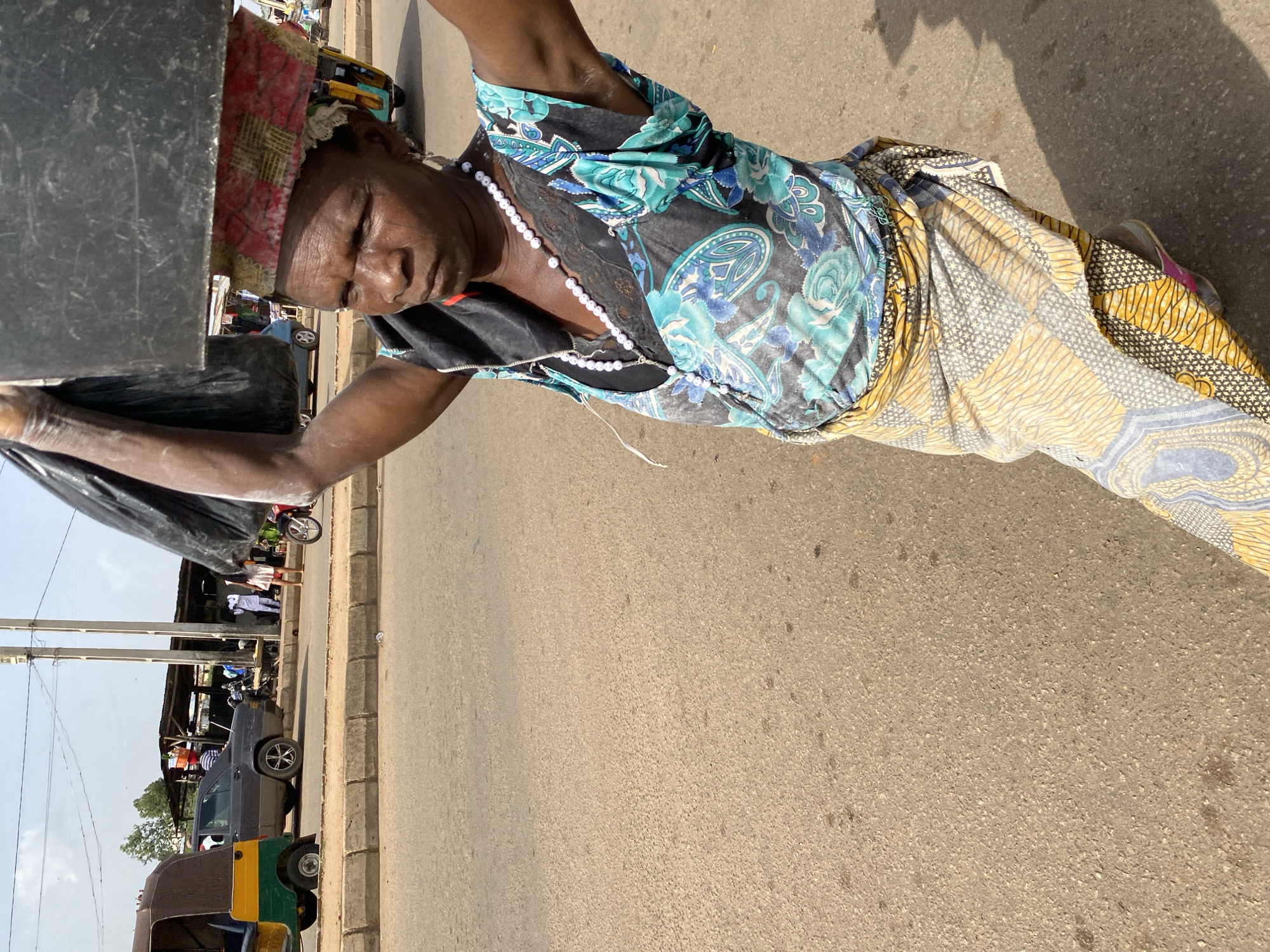 A woman carries a black container on her head as she walks on a paved road in Bukuru, Nigeria. She wears a blue and green patterned blouse and a patterned wrap skirt. She is also wearing a beaded necklace. In the background, there are buildings and vehicles, including a tuk-tuk, a car, and a motorcycle. The weather appears to be sunny.