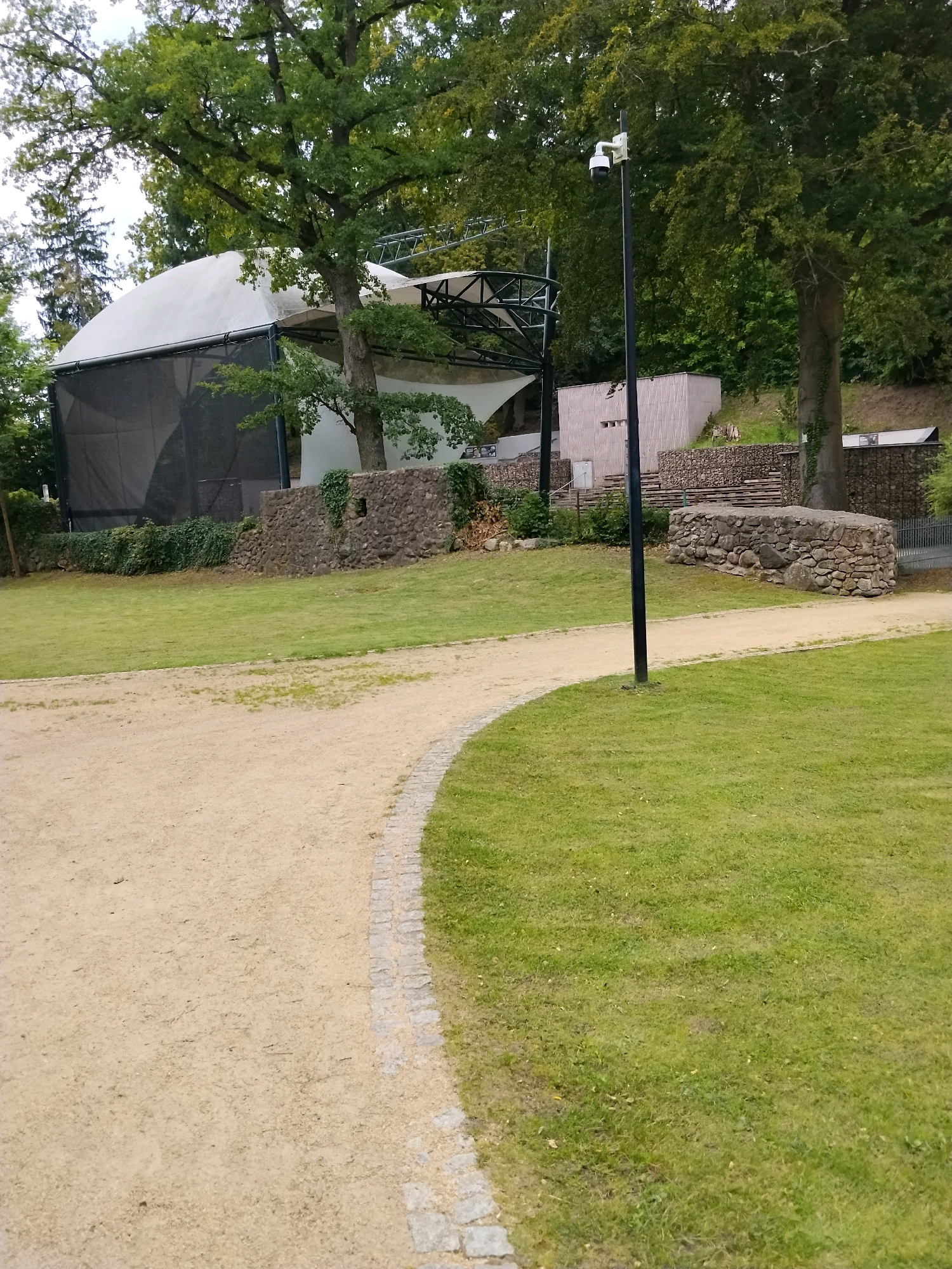 An outdoor, medium-shot, eye-level, slightly downward-angled photograph captures a curved dirt pathway bordered by a stone-and-cobblestone edge on the left, leading into a manicured lawn on the right. The pathway curves away from the viewer toward the left. Beyond the pathway and lawn, a dark-netted, white-covered stage structure is visible, supported by a metal frame. Behind the stage, a large tree with green foliage dominates the background, with a black pole supporting a surveillance camera positioned in front of it. Further behind the stage, a light-colored, corrugated building and a stone wall are discernible, with more trees and foliage receding into the distance. The scene appears to be a public park or outdoor event space in Łagów, Poland, possibly near the Łagów Castle or the amphitheater. There are no people visible in the frame, and the overall atmosphere is calm and serene, suggesting a daytime setting under an overcast sky.