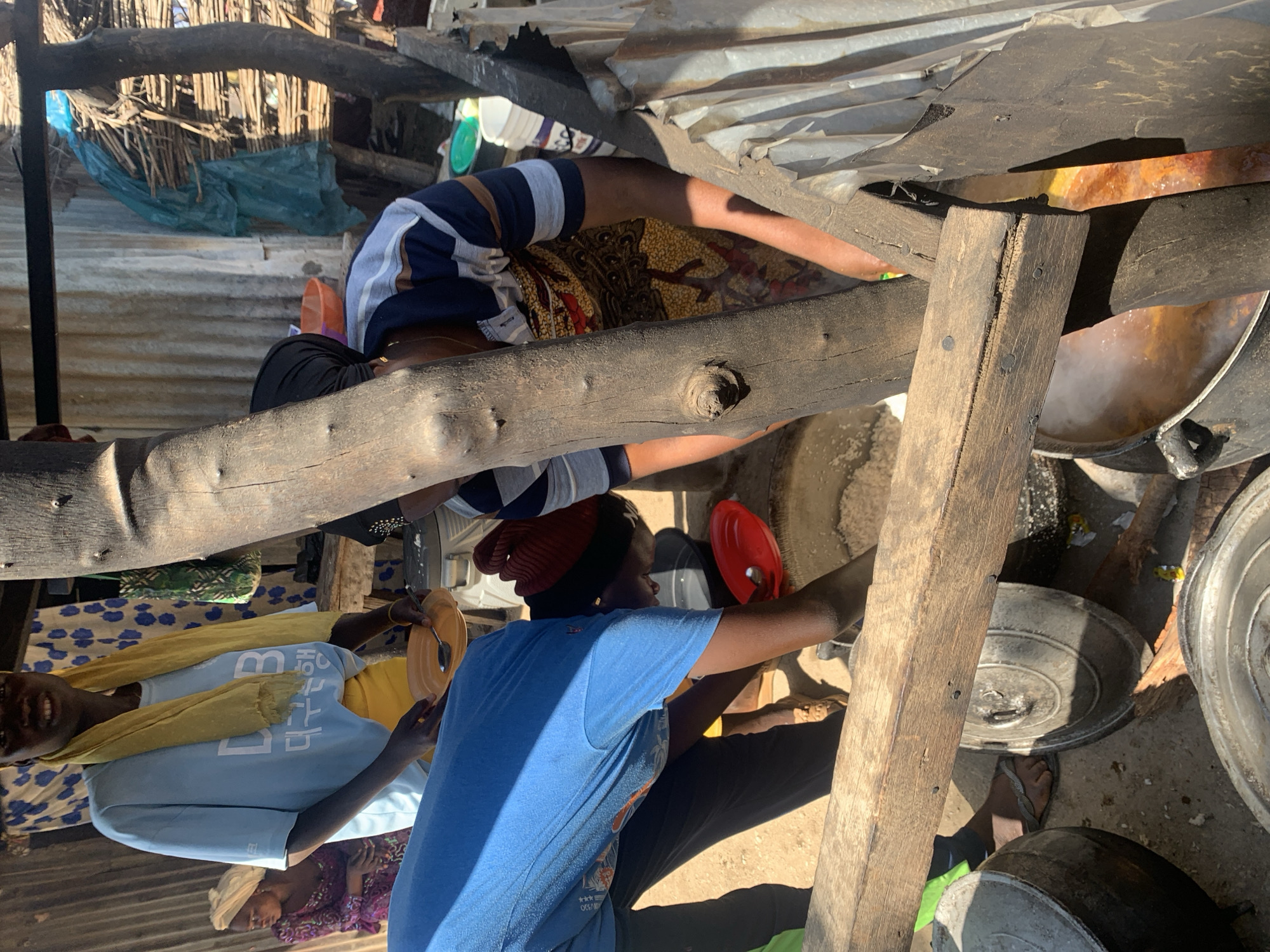 This image depicts an outdoor scene in Nigeria, possibly a market or a communal cooking area, under what appears to be daylight. Several people are engaged in activities related to food preparation or serving.
In the foreground, a person wearing a blue t-shirt with a tropical print and a yellow headscarf is holding a plate and a spoon. They are smiling, suggesting a positive or contented mood. To their left, another person in a blue t-shirt with white text is visible. The text on this shirt appears to be in Korean, with characters that could read "한국" (Hanguk, meaning Korea). Further to the left, a person in a light blue t-shirt and yellow scarf is also present.
In the background, there are elements suggesting a rustic or temporary structure, with corrugated metal roofing and wooden beams. Several large pots and containers are visible, some of which appear to be used for cooking over what might be an open fire, as indicated by steam rising from one of the pots. There are also bundles of reeds or sticks and some plastic sheeting, which could be part of the shelter or materials for the structure.
The overall impression is of a community engaged in daily life, with a focus on food and communal activities. The lighting suggests it is daytime, but the exact time and weather conditions are not definitively clear. The setting seems informal and utilitarian.