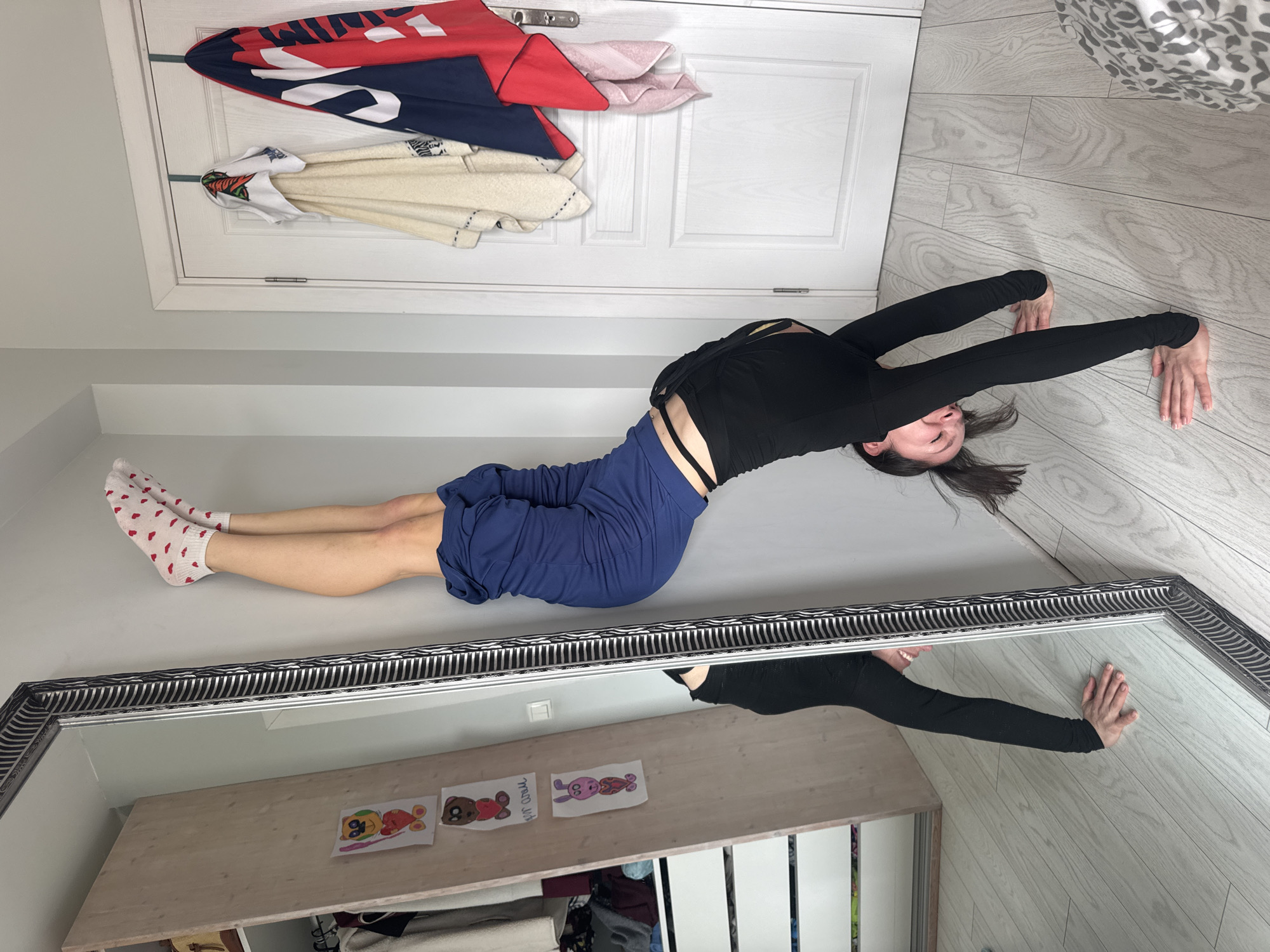 A woman is lying on her stomach, arms outstretched, in what appears to be a room with white walls and light-colored wood-patterned flooring. She is wearing a long-sleeved black top, blue pants, and white socks with red hearts. A mirror reflects the woman and a shelf-like object with drawings on it. A white door with hanging items, including towels and a red and blue garment, is visible. The location cannot be determined as Burgas, Bulgaria from the image.