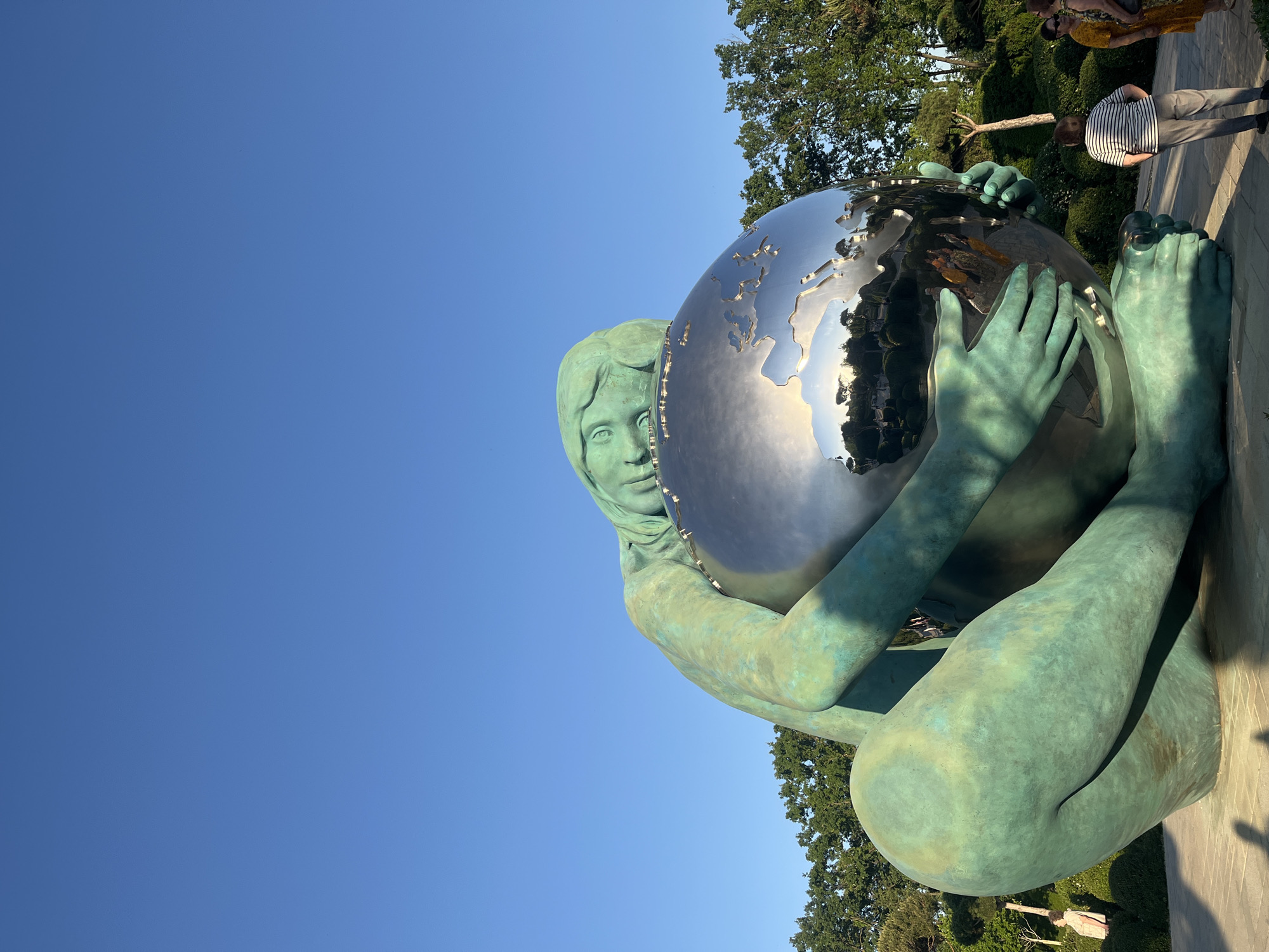 The image displays a large bronze statue of a figure, resembling a young woman, cradling a polished, reflective metallic globe. The statue has a distinctive green patina, suggesting aged bronze, and is depicted in a crouching or seated pose. The figure's face, with a calm expression, is visible peering from behind the left side of the globe, looking towards the viewer. Her hands and feet are prominently featured, holding and supporting the sphere.
The metallic globe, designed to represent the Earth with etched continents, acts as a mirror, reflecting distorted images of the surroundings. These reflections include a bright blue sky, lush green trees, and several people.
In the background, a vast expanse of clear blue sky indicates bright, sunny weather. Green foliage, comprising trees and manicured bushes, suggests a well-maintained park or garden setting. Several individuals are present in the background: some are directly visible on the right side of the statue, casually walking or observing, while others are seen as reflections in the globe. One person is wearing a striped shirt, and another a yellow top.
The scene is illuminated by bright daylight, casting defined shadows, indicating it is either morning or afternoon. The overall impression is one of tranquility in a public outdoor space, showcasing the impressive statue. No visible text is present in the image. The location is Krasnodar, Russia.
