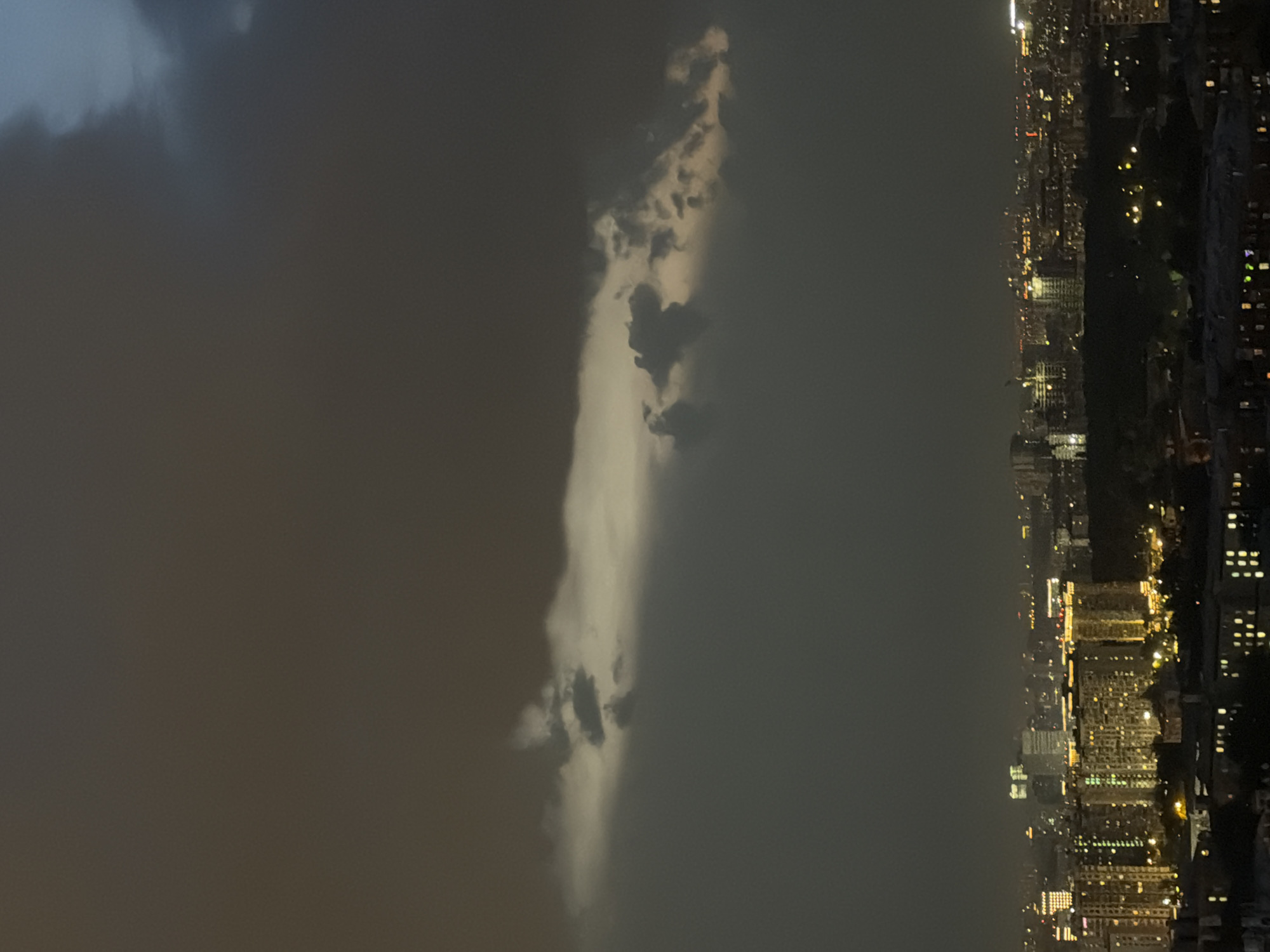 The image captures a nighttime urban scene, possibly from a high vantage point. The right side shows a cityscape with numerous buildings illuminated by artificial lights, indicating residential or commercial activity. The left side is dominated by a dark, cloudy sky with a column of illuminated clouds. The location in the city is Unknown, Unknown.