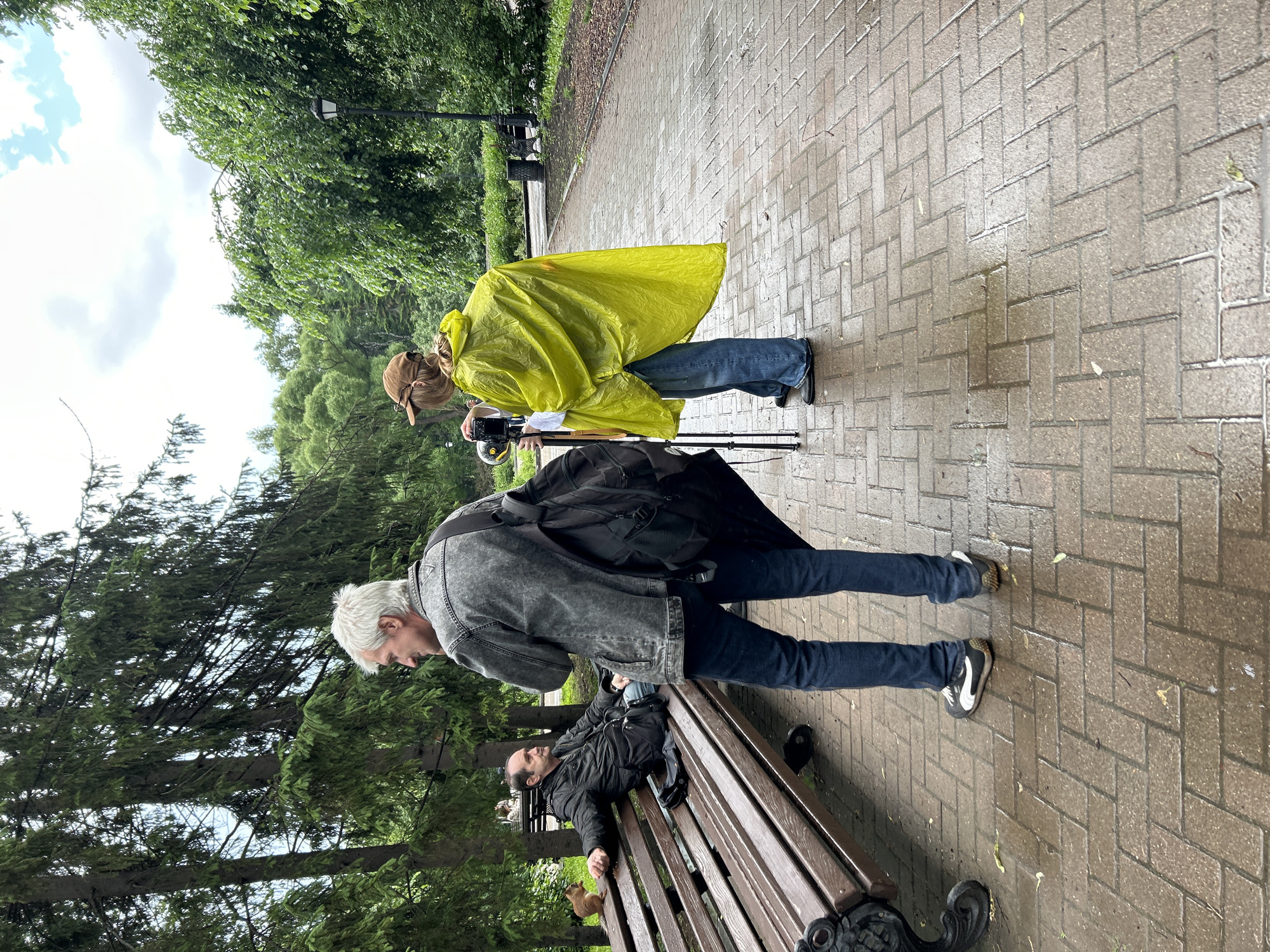 The image depicts an outdoor scene in a park in Yekaterinburg, Russia, following recent rain, evidenced by the wet pavement. A light brown brick-patterned pathway, laid in a herringbone design, extends through the foreground and midground, featuring scattered puddles and leaves. Lush green trees, a mix of conifers and deciduous types, form a dense natural backdrop, filling the upper and background areas under an overcast sky with patches of white clouds.
On the left, an adult male with dark hair and a short beard is seated on a dark brown wooden park bench with black ornate metal legs. He is dressed in a black zippered jacket and dark trousers, looking towards the right, seemingly engaged in a conversation. A small, reddish-brown squirrel is perched on the backrest of the bench to his left, appearing to interact with him or hold something.
In the midground, an adult male with short gray hair, wearing a gray denim jacket with a visible "GUESS" logo on the left chest pocket, dark blue jeans, and black and white Puma sneakers, stands with his back mostly to the viewer. He is turned in profile, facing the man on the bench, and appears to be speaking with him. A large black duffel bag or backpack is slung over his right shoulder.
Further back and to the right, a person wearing a bright yellow rain poncho, blue jeans, and a brown baseball cap stands with their back to the viewer. Their blond hair is tied back in a ponytail. They are operating a camera mounted on a black monopod or tripod, positioned to film or document the interaction between the two men.
In the background, a black ornate lamppost is visible on the right side of the path, with another park bench and a black trash can further along the pathway. The overall lighting is diffused, consistent with an overcast day.