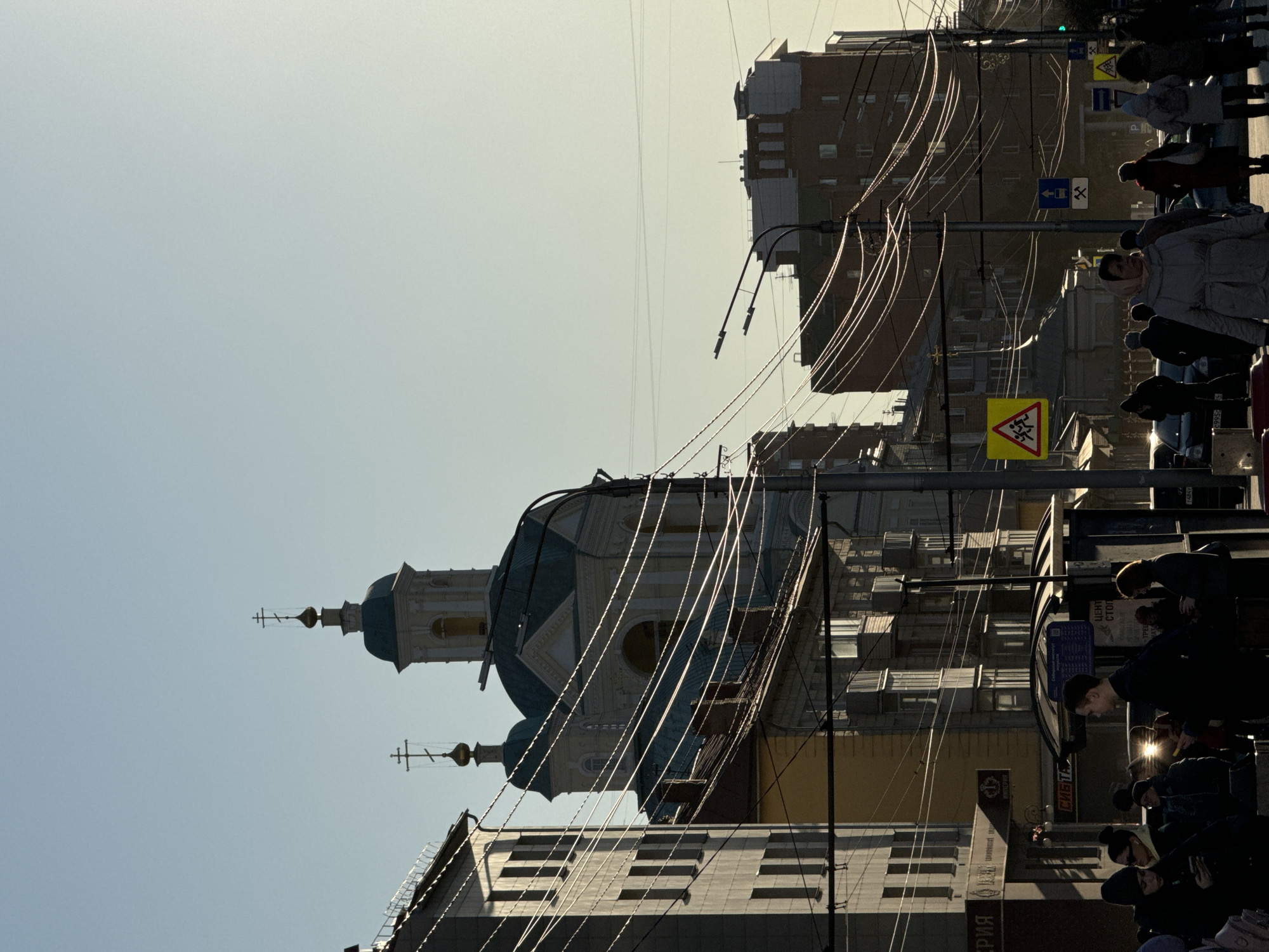 Captured in Krasnoyarsk, Russia, the image shows an urban street scene. A church with a distinctive blue dome and cross-topped spire stands amidst buildings. Utility lines traverse the sky, adding to the cityscape complexity. People are present on the street, some gathered near what appears to be a bus stop. Street signs are visible, indicating a pedestrian crossing. The lighting suggests it is daytime, with shadows cast by the buildings.