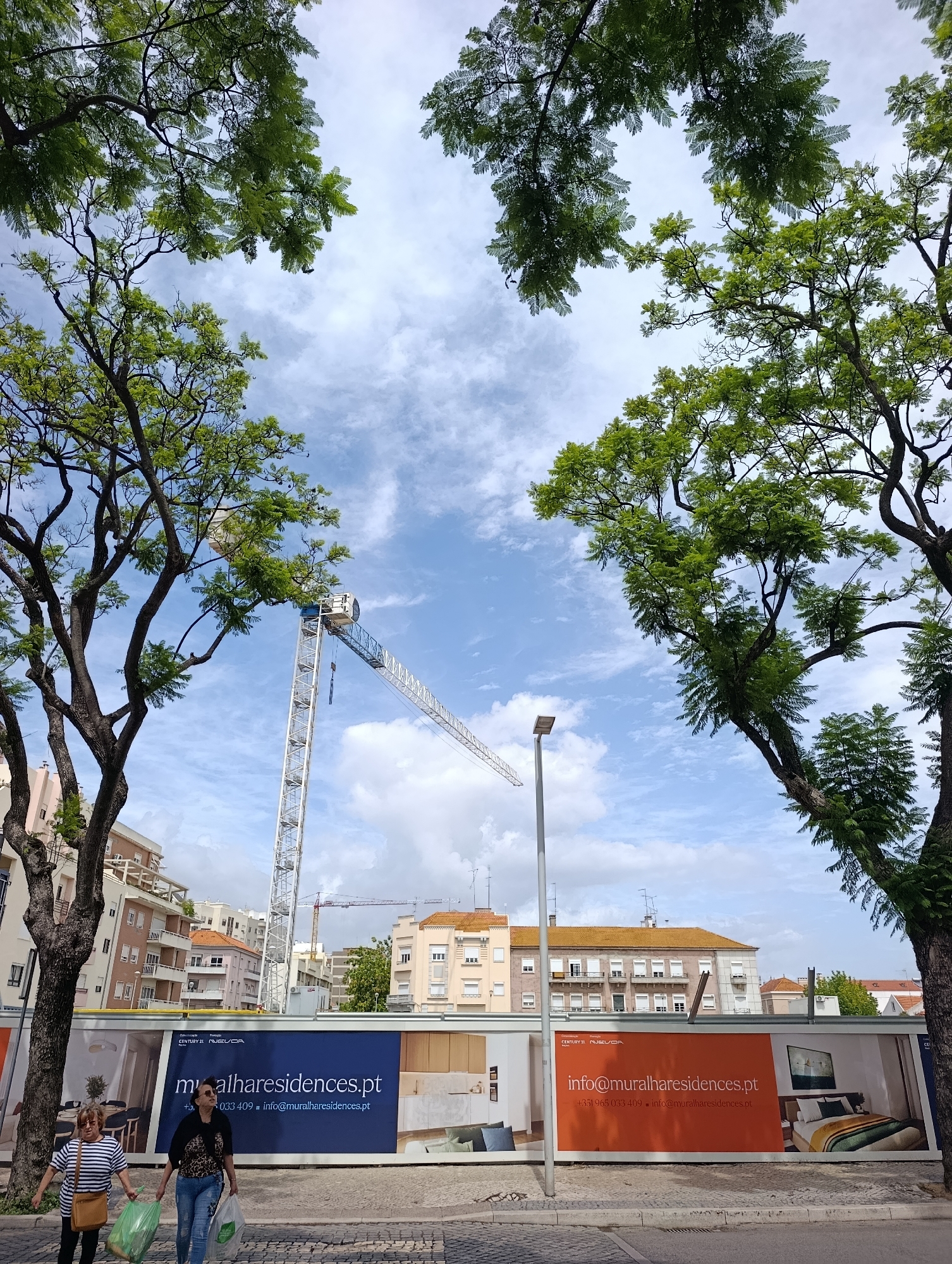 The image shows a street scene in Setúbal, Portugal. The foreground features two women walking on a sidewalk. The woman on the left wears a striped shirt and a light brown shoulder bag, carrying two green plastic bags. The woman on the right wears a leopard-print top and jeans, carrying a clear plastic bag. Behind them, a construction fence displays advertisements for "muralharesidences.pt". In the background, buildings and a construction crane are visible against a cloudy sky. Trees with green foliage frame the scene from above. A street lamp stands to the left of the fence.