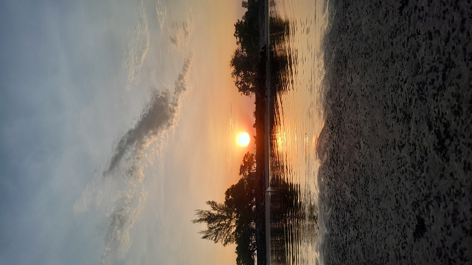 The image depicts a coastal or estuarine scene at sunset or sunrise in Lhokseumawe, Indonesia. The sky transitions from a pale blue-grey at the top to vibrant orange and yellow hues at the horizon, with scattered cirrus and cumulus clouds present. The sun, a bright orange-yellow orb, is positioned low on the horizon, slightly left of the image's vertical center. Its light creates a prominent, shimmering reflection across the calm body of water occupying the middle ground.
A dark silhouette of a distant shoreline extends across the horizon, featuring dense foliage, including tall, slender pine-like trees on the left and other varied tree species. Rocky outcrops are discernible along this distant waterline. A single individual is visible in the shallow water near the distant shore, positioned left of the sun's reflection. This person appears to be engaged in fishing, with what resembles a net partially submerged in the water. The immediate foreground consists of dark, textured sand or mud flats, likely exposed by low tide, displaying an uneven, pitted surface extending from the bottom of the frame to the water's edge.