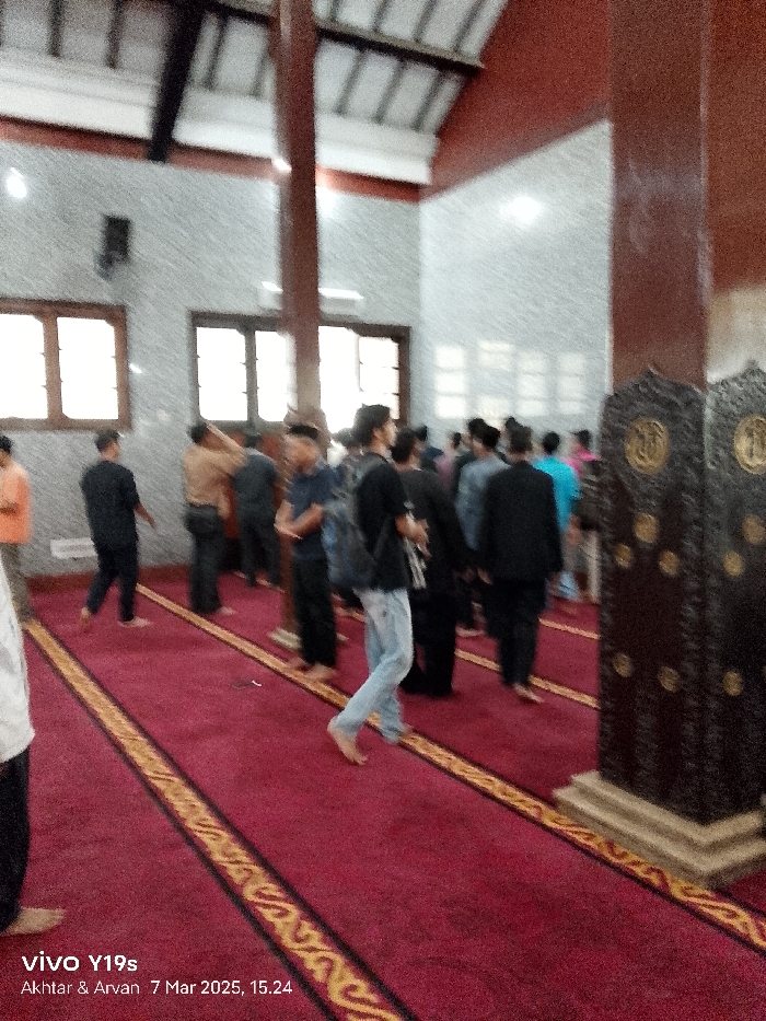 The image shows a mosque in Indramayu, Indonesia, located at coordinates -6.327 latitude and 108.322 longitude. The mosque is filled with men, all wearing clothing common in the Muslim faith. Some of the men are standing and talking, while others are sitting on the carpeted floor. The men are gathered around two large wooden pillars, and there is a red rug covering the floor. The room is lit by natural light coming through large windows. There is a large wooden door at the far end of the room, which is also covered in a red rug. The image appears to have been captured by a mobile device and features a date stamp of 7 March 2025, 15:24.