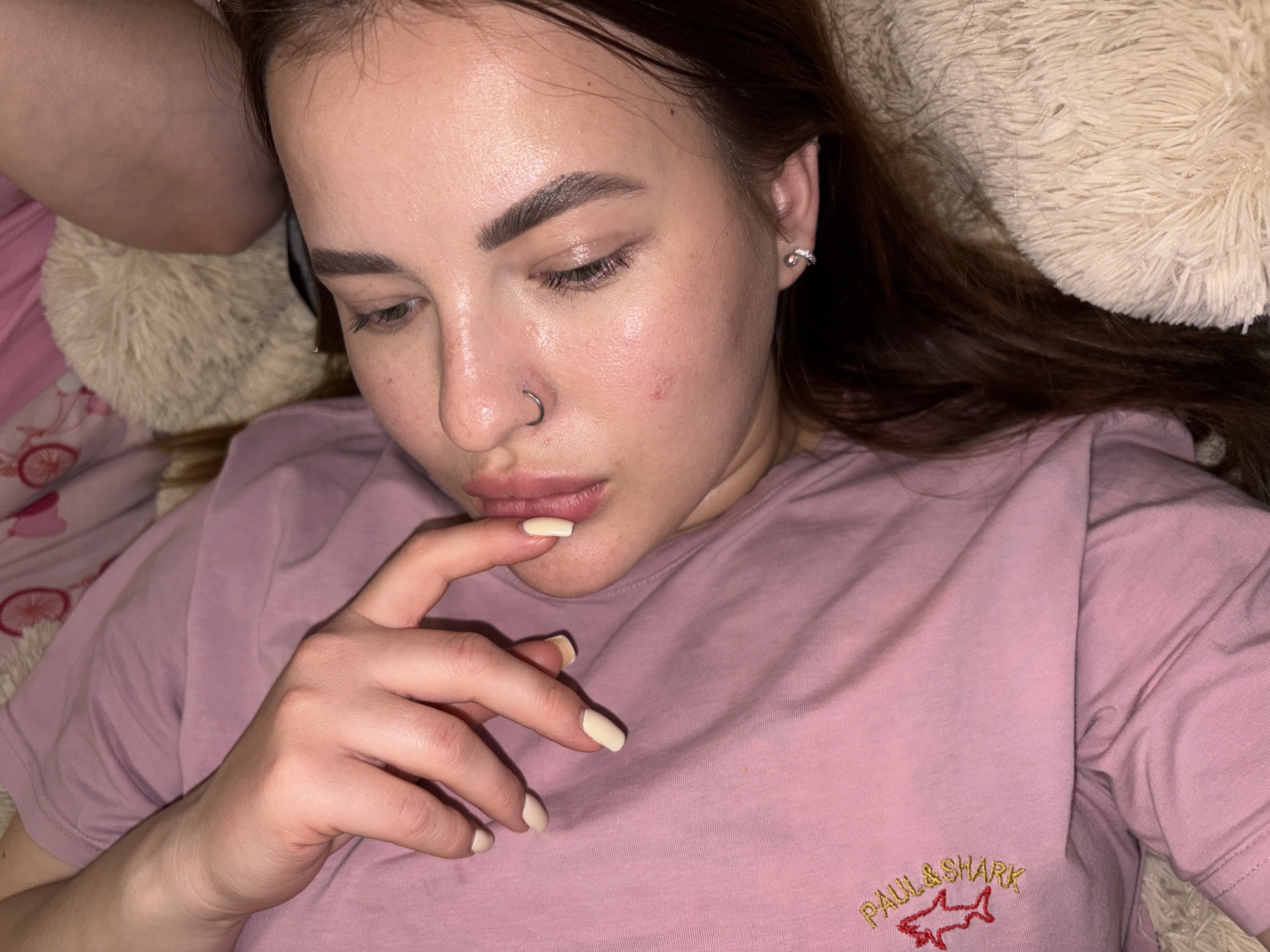 The image captures a young woman lying down in an indoor setting, likely a bed or sofa, with soft textures surrounding her. She is seen from an overhead perspective, with her gaze directed downwards and to her right, giving her expression a pensive or relaxed quality.
Her dark brown hair is spread out, blending with a fluffy cream or beige pillow or blanket that supports her head. Her skin appears natural, with a slight sheen on her forehead and nose, and some minor redness visible on her right cheek. She has prominent eyebrows and visible eyelashes. A silver hoop nose ring is worn on her right nostril, and a small silver hoop earring with tiny stones is visible in her right earlobe.
She is wearing a light pink short-sleeved t-shirt. Embroidered on the front of the shirt, in yellow thread, is the text "PAUL & SHARK," beneath which is a distinctive red shark logo. Her right hand is raised, with her index finger gently touching her lower lip, contributing to her thoughtful posture. Her fingernails are manicured with a light, possibly off-white or beige, polish. To the far left, another pink fabric, possibly bedding, with a subtle bicycle pattern, is visible. The lighting is soft and ambient, indicating an indoor environment where she appears to be resting or contemplating.