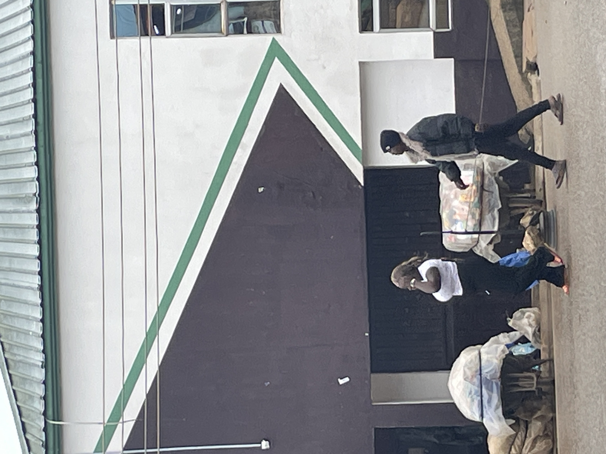 In Bukuru, Nigeria, two individuals are observed walking on a paved surface in front of a building with a dark brown and white facade featuring a prominent green diagonal stripe. On the left, a woman with dark, medium-length hair is walking and speaking on a cell phone. She is wearing a white t-shirt and black pants. To her right, a man wearing a dark beanie, a dark jacket with a furry collar, and dark pants is walking. Both individuals appear to be walking away from the camera, towards the right side of the frame. In the foreground, to the left of the woman, are two carts laden with goods covered in white plastic sheeting and burlap sacks. To the right of the man, a section of a sloping walkway or ramp is visible. The building behind them has a corrugated metal roof and multiple windows. The overall scene suggests a street-level view in an urban environment.