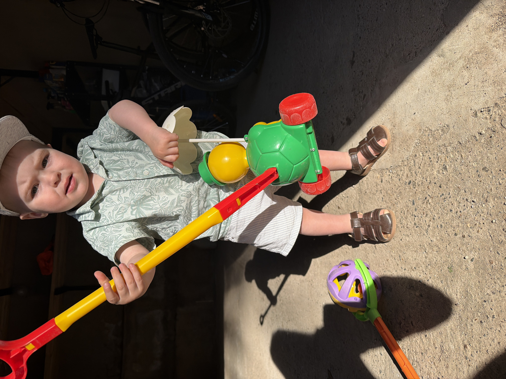 A young child, likely a toddler, stands on a concrete surface. The child wears a light-colored hat, a patterned green shirt, and striped white shorts with brown sandals. The child is holding a yellow and red plastic toy shovel and a green and yellow toy. Near the child's feet, there is a purple, yellow, and green toy ball on a stick. In the background, a bicycle is visible in a dimly lit area. The scene is partly in sunlight, casting distinct shadows of the toys and the child. The location is potentially Votkinsk, Russia, but there is no confirmation from the image.
