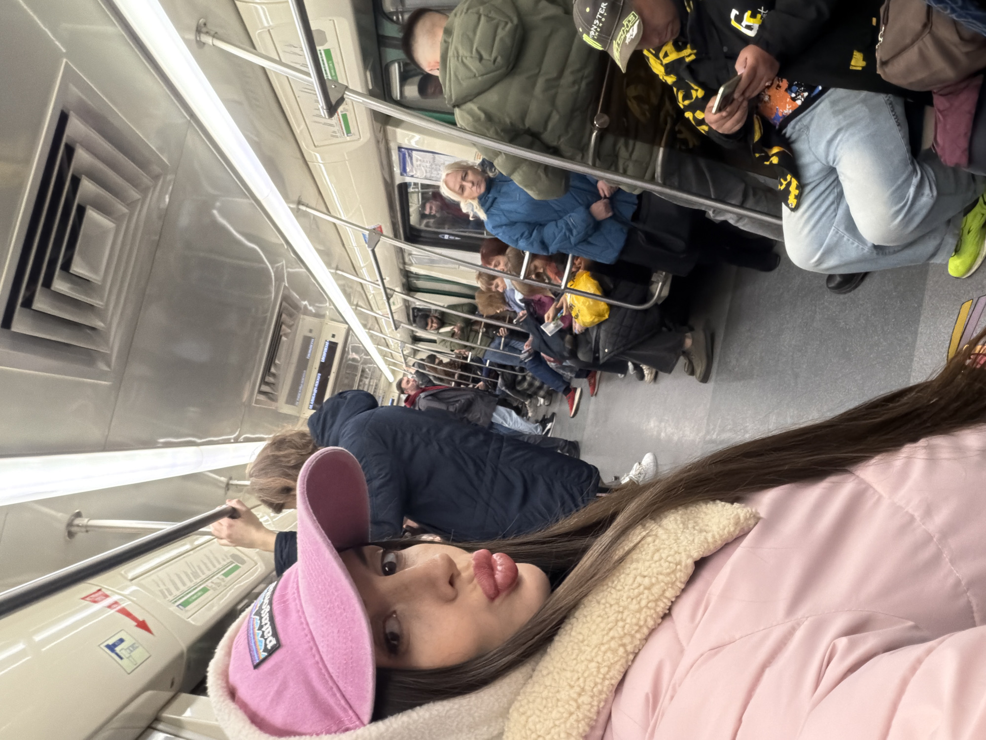 The image shows the interior of a subway car, possibly in Saint Petersburg, Russia, as indicated by the style of the subway car and its features.
The subway car is crowded with numerous passengers, including a woman in the foreground wearing a pink jacket, a pink baseball cap, and long brown hair. She is looking at the camera with a pout.
Several other passengers are visible in the background. Many of them are standing and holding onto poles. Most are dressed in winter clothing, including jackets and coats. Some appear to be looking at their phones or other devices.
The interior of the subway car is mostly stainless steel. The overhead lighting consists of long fluorescent lights. Subway information signs are visible along the walls. The floor is gray. Overall, the scene suggests a typical urban commute.