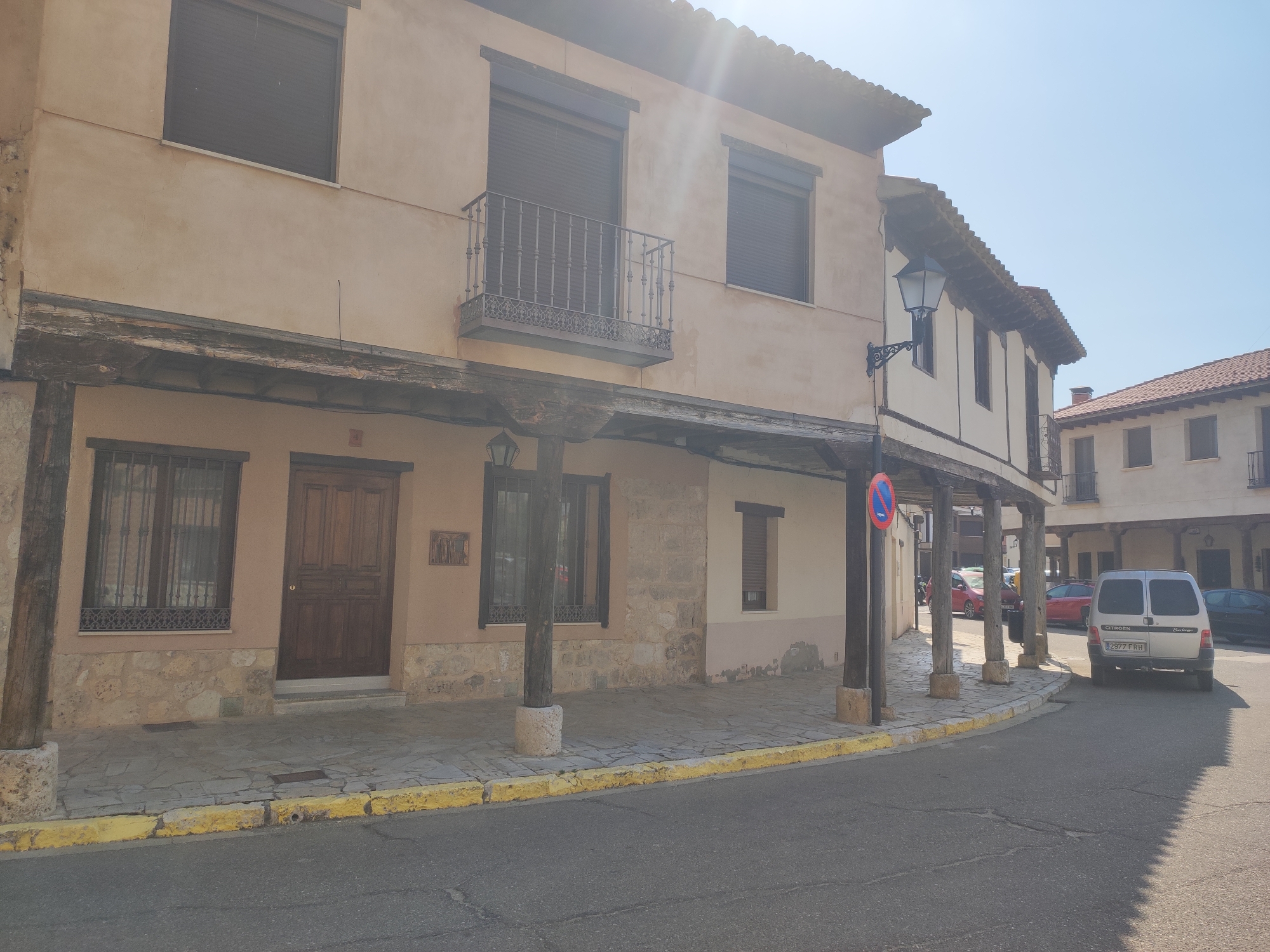 The image depicts a street scene in Ampudia, Spain, characterized by traditional architecture. In the foreground, a paved road curves to the right, with a yellow-painted curb bordering a stone-tiled sidewalk. The sidewalk is lined with wooden pillars supporting an overhang of an old building. The building's facade is a light beige stucco with stone accents at the base. It features several windows with dark shutters and a balcony with an ornate metal railing on the upper floor. A dark wooden door is visible on the ground floor, along with a barred window.
Further down the street, other buildings with similar architectural styles are visible, including terracotta tiled roofs and wooden beams. Several vehicles are parked along the street, including a silver Citroen van with the license plate "2977 FRH", a red car, and a black car. A street lamp with a black ornate fixture is attached to one of the buildings. A "no parking" sign is posted on a pole. The sky is clear blue, indicating a sunny day. The sun casts a lens flare in the upper right portion of the image.