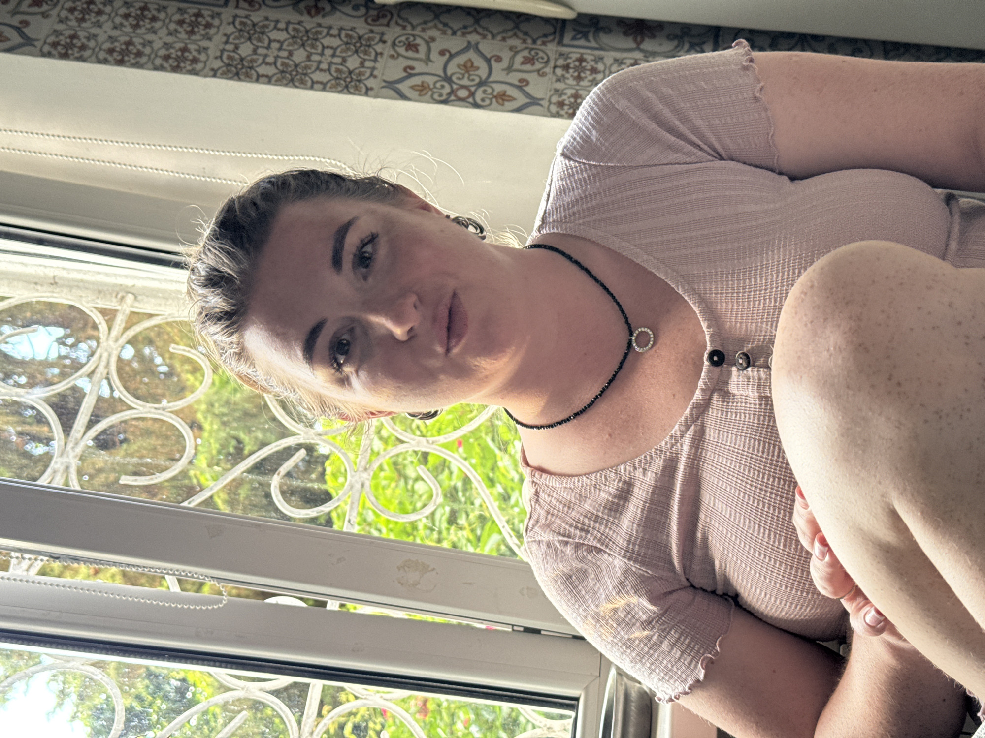 A woman is seated indoors, with a window providing a view of green foliage and decorative metalwork. She is wearing a light pink, short-sleeved, ribbed top with buttons and a black necklace with a circular pendant. Her left arm is resting on her bent left leg. Her hair is pulled back, and she is looking towards the camera with her mouth slightly open. The background features a patterned curtain and a white wall.