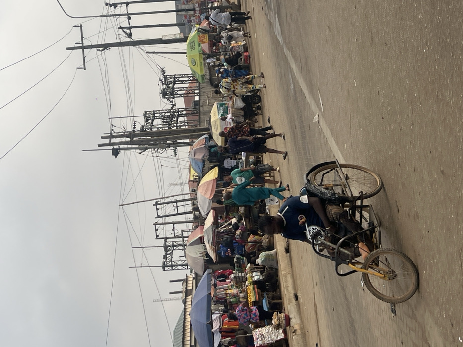 The image depicts a bustling street scene in Egan, Nigeria, under an overcast sky. In the immediate foreground, a dark-skinned male in a dark blue short-sleeved shirt and patterned trousers operates a self-propelled, three-wheeled mobility device on an asphalt road. The device features two large rear wheels and a smaller front wheel with a yellow fork. To the left, a vibrant street market occupies the roadside, characterized by multiple vendor stalls shaded by diverse umbrellas (blue, striped, green, yellow). Various goods, including produce and clothing, are displayed on tables and benches. Numerous pedestrians traverse the market area and the adjacent road. Some individuals are seen walking, while one person further back pushes a cart. The background is dominated by a complex network of utility poles, overhead electrical wires, and multiple large electrical transformers. Low-rise buildings with corrugated roofs are partially visible behind the market stalls.