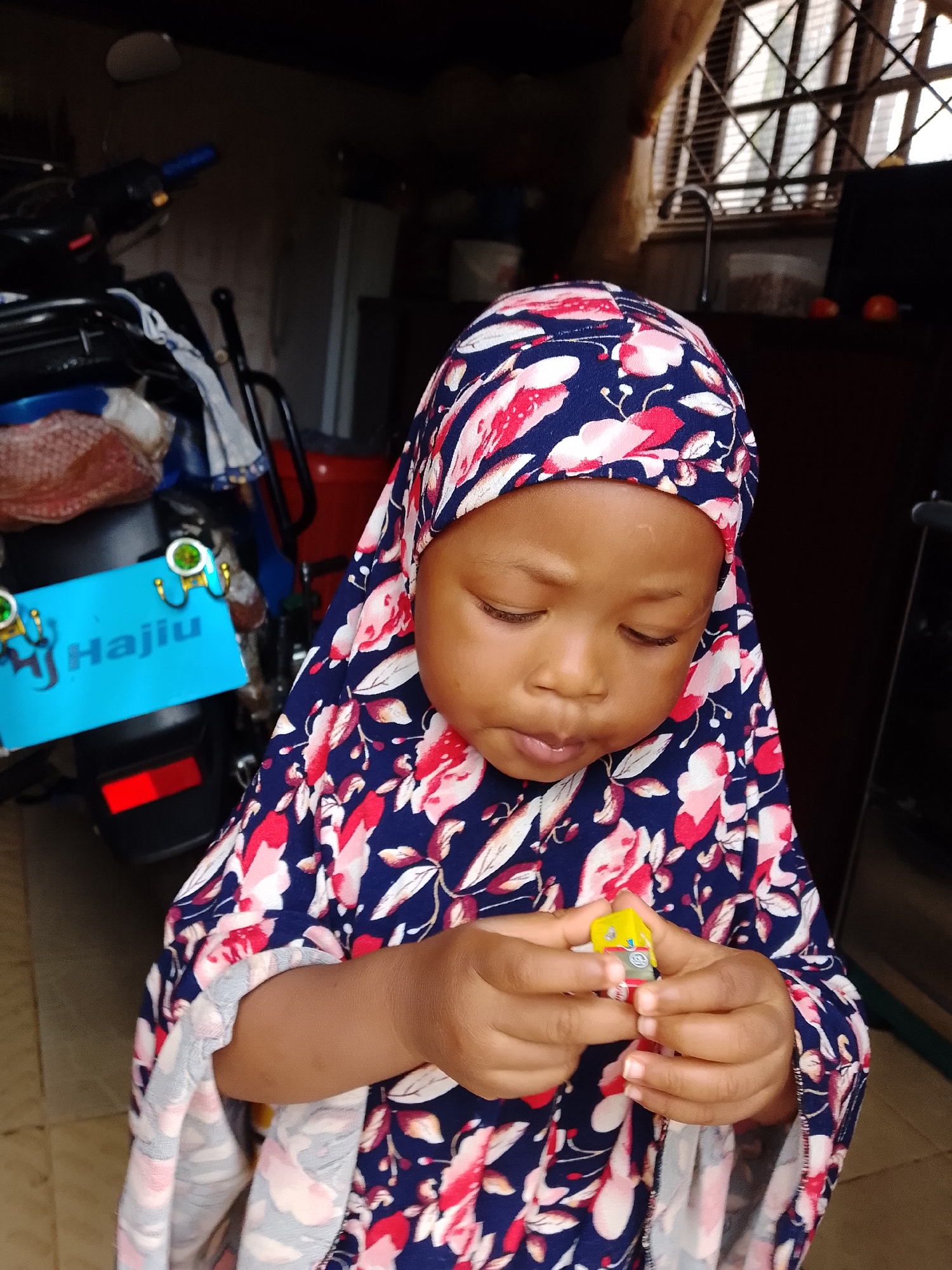 A close-up, waist-up, medium shot shows a young Black child wearing a navy blue hijab with a pink and white floral pattern. The child is looking down at their hands, which are holding a small, yellow, rectangular object with red and silver accents. The child's skin is a medium brown, and their eyes are closed. The child's left hand is visible, showing fingernails. The child's right arm is also visible, with a bit of the floral fabric of their garment. The background is dark and out of focus, but appears to be an indoor or semi-enclosed space. A blue sign with white text that reads "Hajjiu" is visible on the left side of the image, attached to what appears to be the back of a motorcycle. The sign has two green circular embellishments. The child's clothing is a navy blue fabric with a repeating pink and white floral pattern. The image appears to have been taken in Jos, Nigeria.