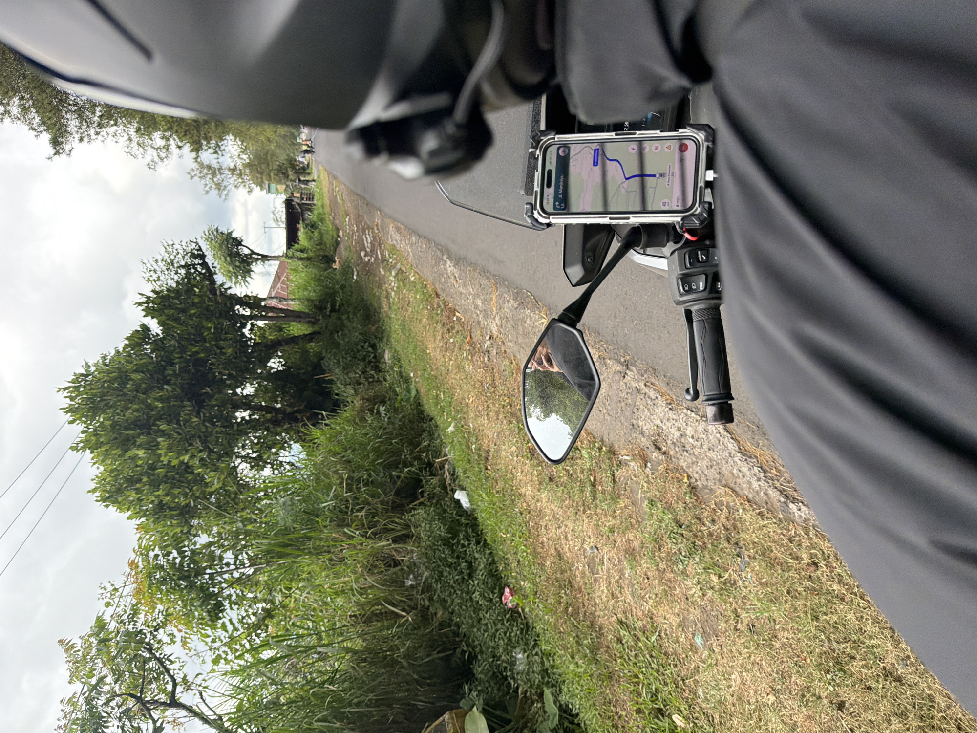 The image depicts a view from the perspective of a person riding a motorcycle in Kintamani, Indonesia. The rider is wearing a black helmet and a black jacket. A smartphone mounted on the handlebars displays a navigation app showing a route with the destination "Jl. Manukiyu". The road ahead is paved and appears to be a rural or semi-urban area. To the left of the road, there is lush green vegetation, tall grasses, and trees. Some discarded plastic waste is visible in the grassy area. The sky is overcast. In the distance, a few other motorcycles and structures can be seen. The motorcycle's side mirror reflects the rider and the surrounding environment.