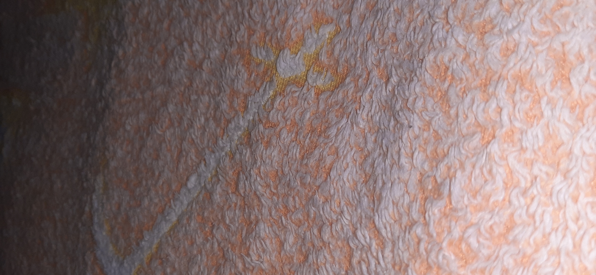 This is a close-up, angled shot of a textured surface, likely a blanket or rug, with a pattern resembling a stylized sword. The texture is plush and uneven, with visible fibers. The dominant colors are off-white and a peachy-orange, with hints of yellow in the patterned area. The lighting is dim, creating soft shadows and highlighting the texture. The image appears to be taken indoors. There are no people or overt actions depicted. The location in Yessentukskaya, Russia is not visually discernible from the image itself.