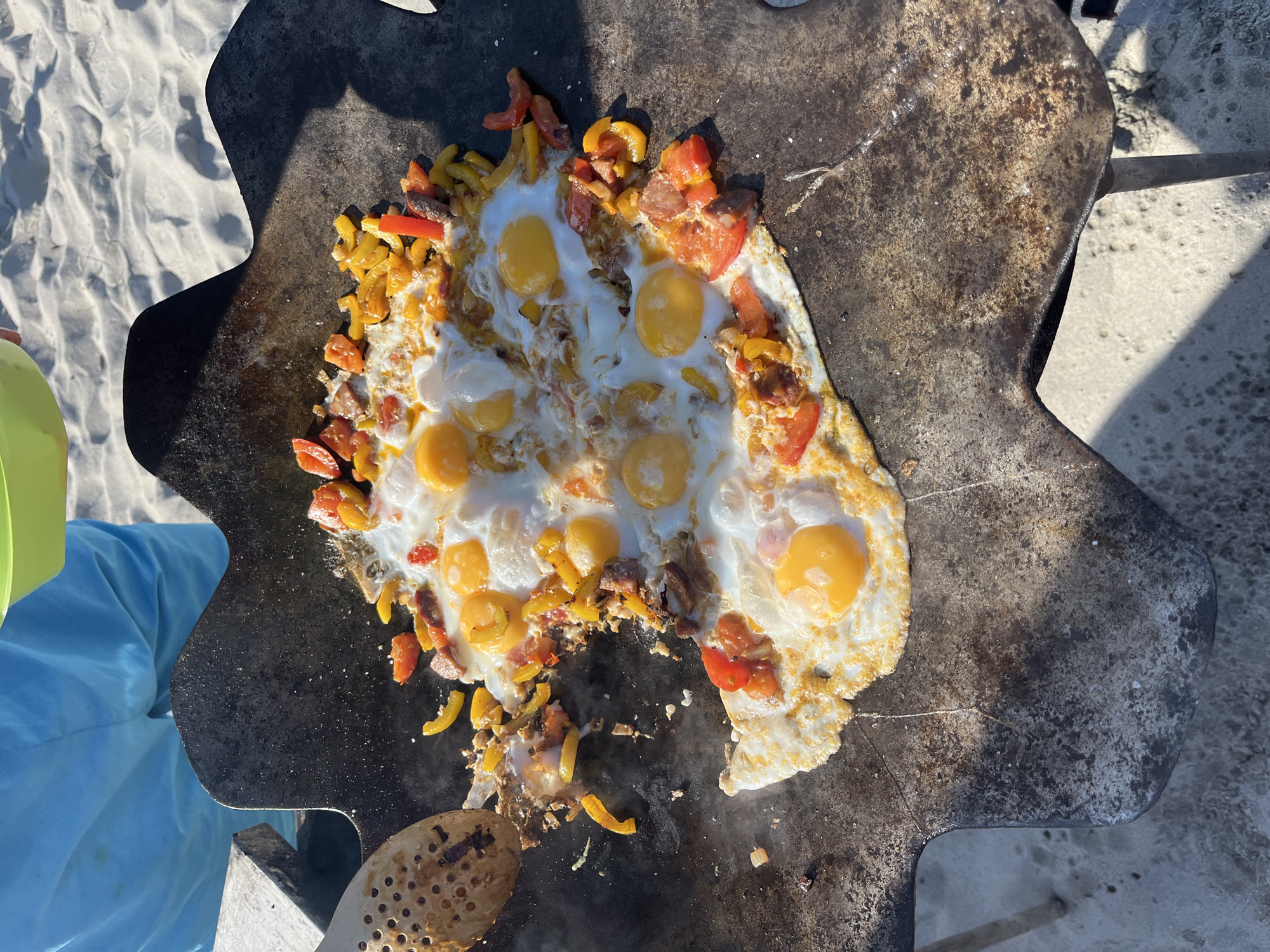 This is a high-angle, eye-level shot of fried eggs with vegetables and meat being cooked on a flat, irregular-shaped griddle over sand. The griddle appears to be a modified shovel head. The food consists of several fried eggs, some with runny yolks, mixed with chopped red tomatoes, yellow and red bell peppers, and pieces of sausage or other meat. The griddle is dark and appears to be made of cast iron, showing signs of use and some rust. To the left of the griddle, a slotted wooden spatula is visible, positioned as if in use. In the background, a person wearing bright blue shorts is partially visible, holding a lime green bowl. The ground is covered in light-colored sand, suggesting an outdoor location, possibly a beach. The lighting indicates a sunny day.