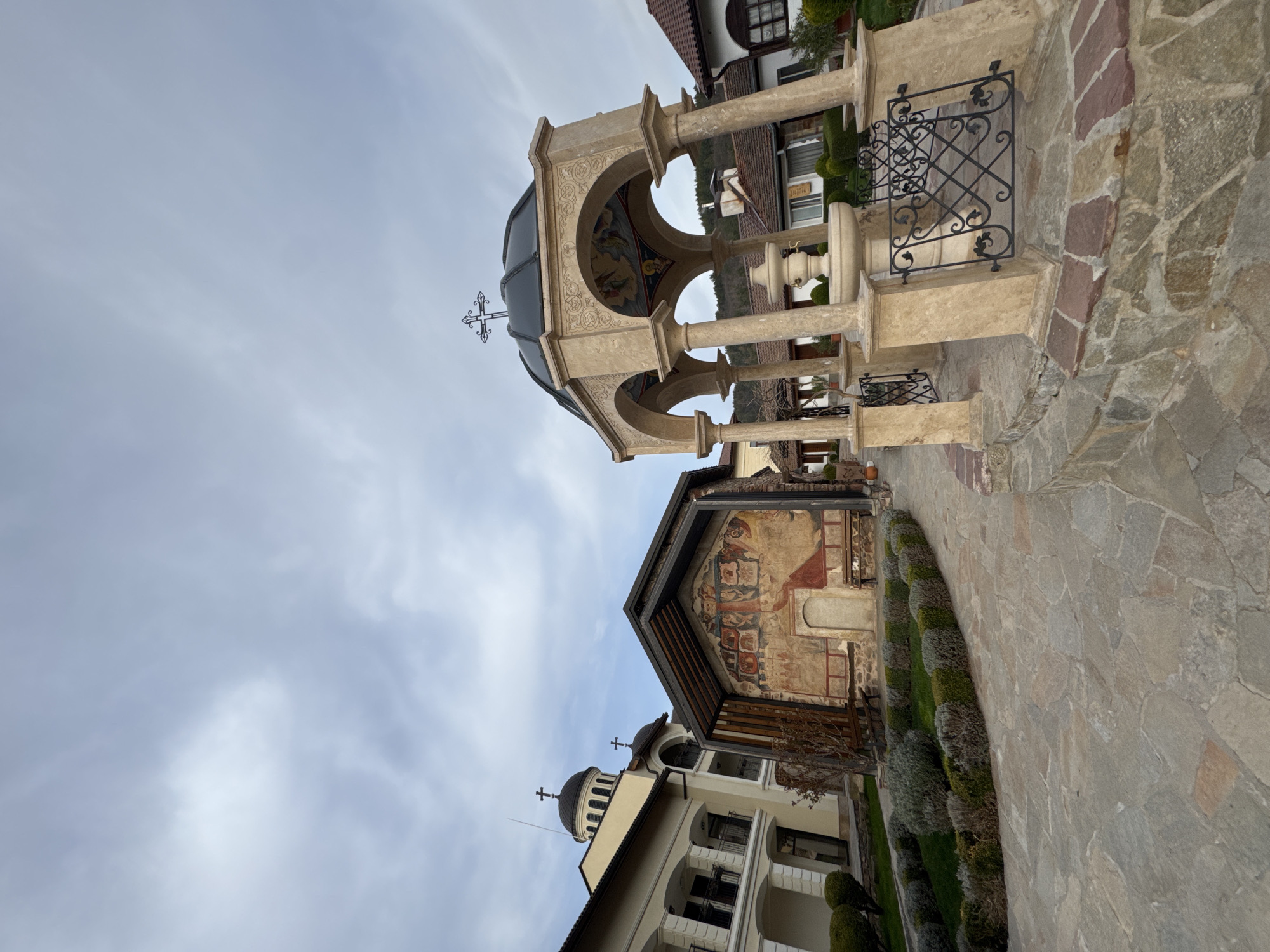 The image captures an outdoor scene at the Boyana Church in Sofia, Bulgaria. A stone pathway leads to an ornate stone structure with a dome topped by a cross, flanked by columns and ironwork. Behind it is a building with exterior frescoes. In the background, additional structures are visible, with a cloudy sky overhead.
