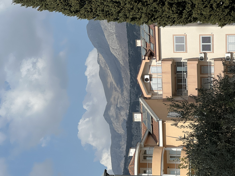 The image displays a daytime scene in Budva, Montenegro, under a partly cloudy sky. The foreground features dense green foliage, with a tall, dark green coniferous tree vertically bordering the right side of the frame. In the midground, three multi-story residential buildings are visible. The leftmost building, partially obscured, exhibits a light peach facade with a rounded balcony and a red-tiled roof. The central structure, also light peach, has a red-tiled roof equipped with multiple solar thermal panels and a single visible balcony. The building on the right, distinguished by a light beige facade, presents multiple balconies with white railings, white louvered window shutters (some open, some closed), and several externally mounted white air conditioning units. Its red-tiled roof also features solar thermal panels. In the background, a range of grayish-brown mountains with sparse vegetation stretches across the horizon. The sky above is blue with scattered white cumulus clouds. No individuals, vehicles, or specific activities are discernible within the scene.