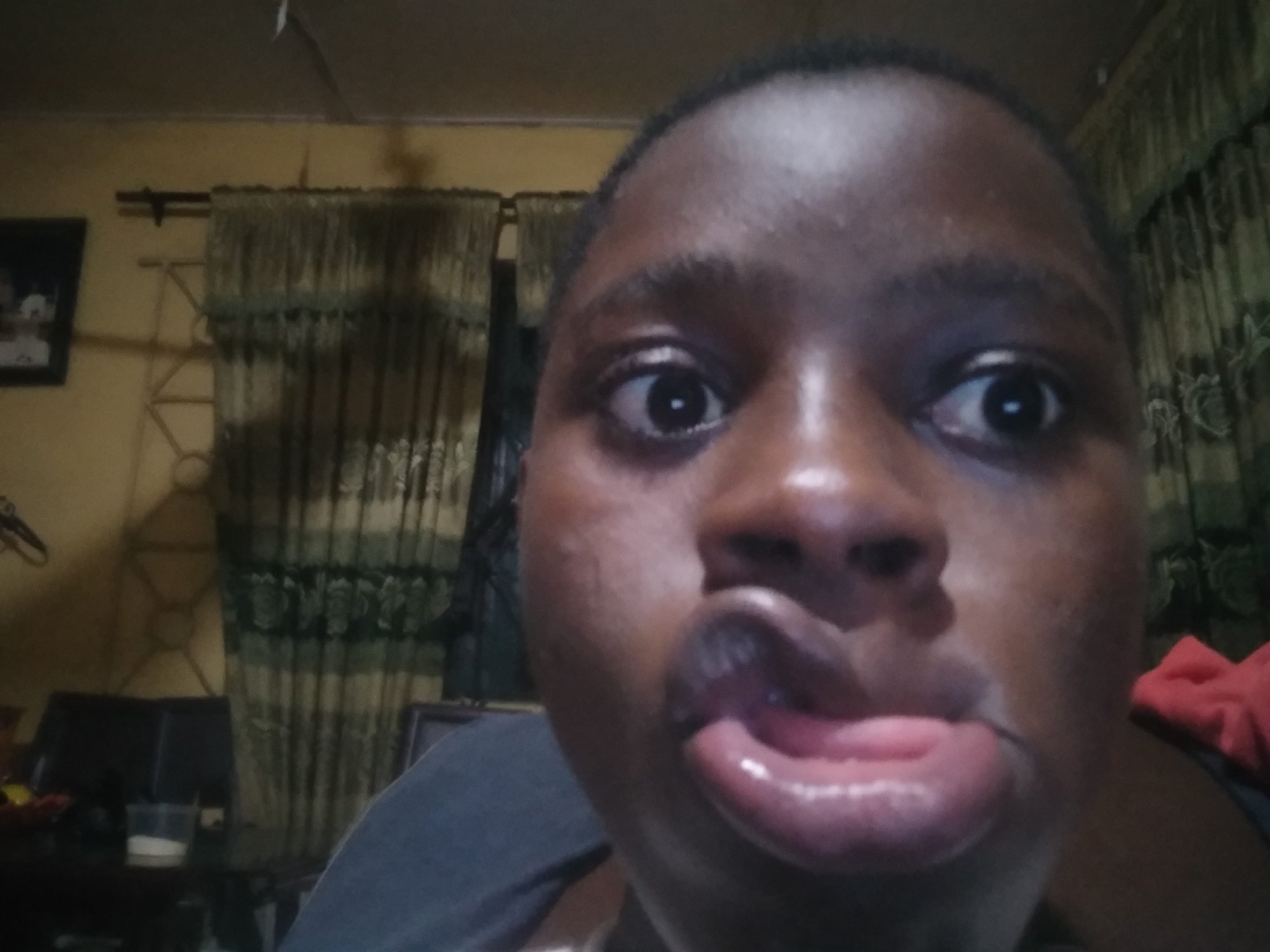 A close-up, poorly lit photo features a dark-skinned person with wide eyes and their mouth contorted into an odd expression. The person is indoors, with a curtain visible in the background. A picture frame and other indistinct objects are partially visible on a table to the left. The location is Unknown, Unknown.