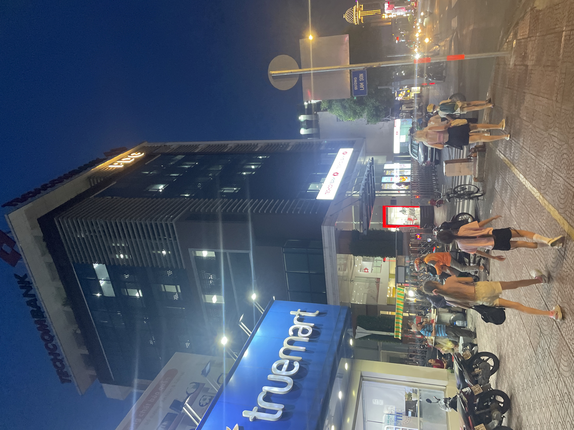 This is a nighttime, eye-level photograph of a street scene in Thủ Đức, Vietnam, featuring the Techcombank building and the Truemart store. The Techcombank building is a tall, modern structure with illuminated signage at the top. Adjacent to it, the Truemart store is visible, its blue signage brightly lit. In the foreground, several people are walking on the sidewalk. Two young girls in casual attire are walking away from the camera, one with a soccer ball at her feet. Behind them, a woman in a pink top and black skirt is also walking away. Other individuals are present in the background, some on motorcycles and others near a food stall. Traffic lights and streetlights illuminate the scene, reflecting on the wet pavement. A street sign indicates "DUONG LAM SON." The overall atmosphere is busy and urban, typical of a commercial street in the evening.