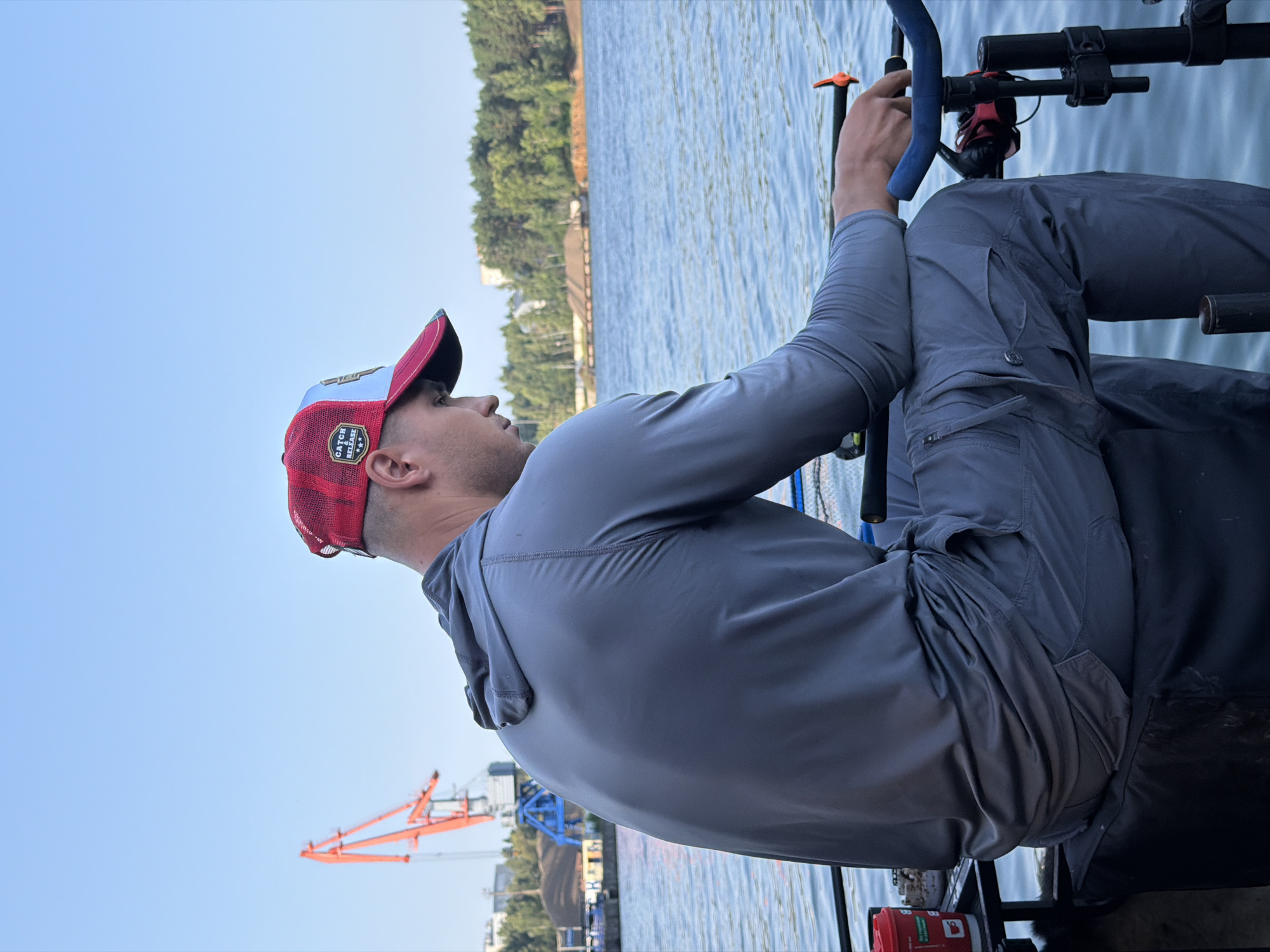 The image depicts a man sitting on a black seat with a gray hoodie and pants, wearing a red and white baseball cap. He is looking down at his hands, which are resting on a fishing rod holder. The setting appears to be a waterfront area, possibly a harbor, with a large orange crane visible in the background. The man is positioned facing the water, which reflects the clear blue sky. The overall scene suggests a calm and focused moment, possibly related to fishing. The location is the city of Chaikovsky, Russia, near the Kama River.