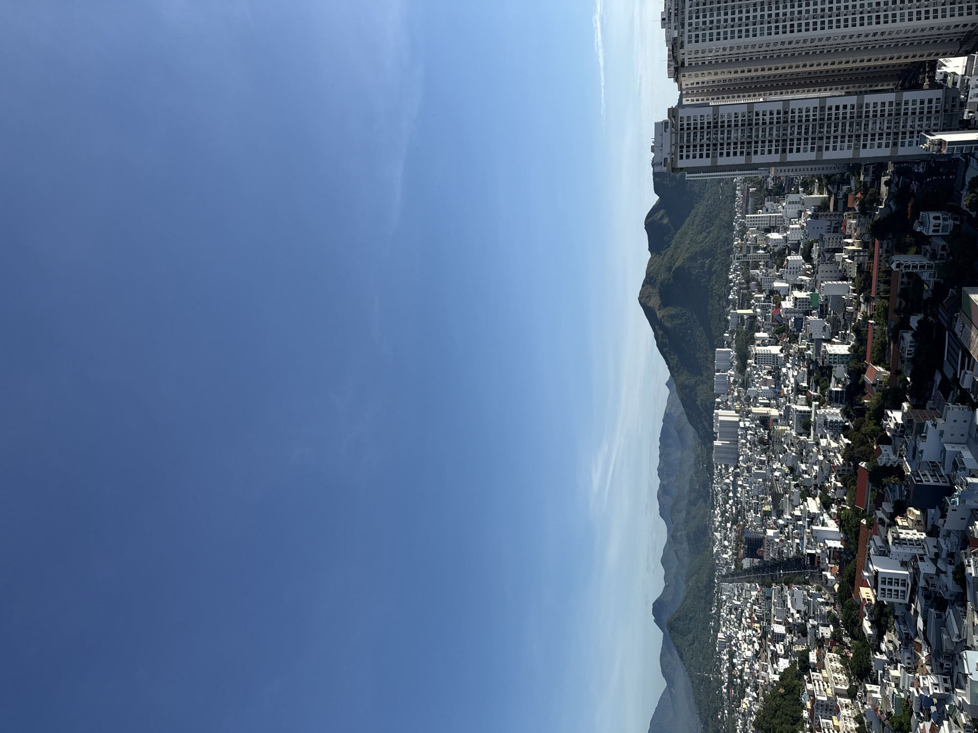 The image shows an elevated, panoramic view of the city of Nha Trang, Vietnam, under a clear blue sky. The foreground is dominated by the tops of several tall, modern apartment buildings, two of which are particularly prominent on the right side of the frame. These buildings feature numerous windows and balconies.
Beyond the apartment buildings, the city stretches out with a dense collection of smaller buildings, a mix of residential and commercial structures, characterized by white and light-colored facades. A long, straight road, possibly a main artery of the city, is visible cutting through the urban landscape.
In the background, a range of lush, green mountains forms a dramatic backdrop to the city. The slopes of these mountains are covered in vegetation, and they appear to be a natural preserve or parkland. The lighting suggests it is daytime, with the sun illuminating the scene from the side, casting some shadows within the city. There are no people or specific actions visible in this image; it is a static, expansive cityscape.
