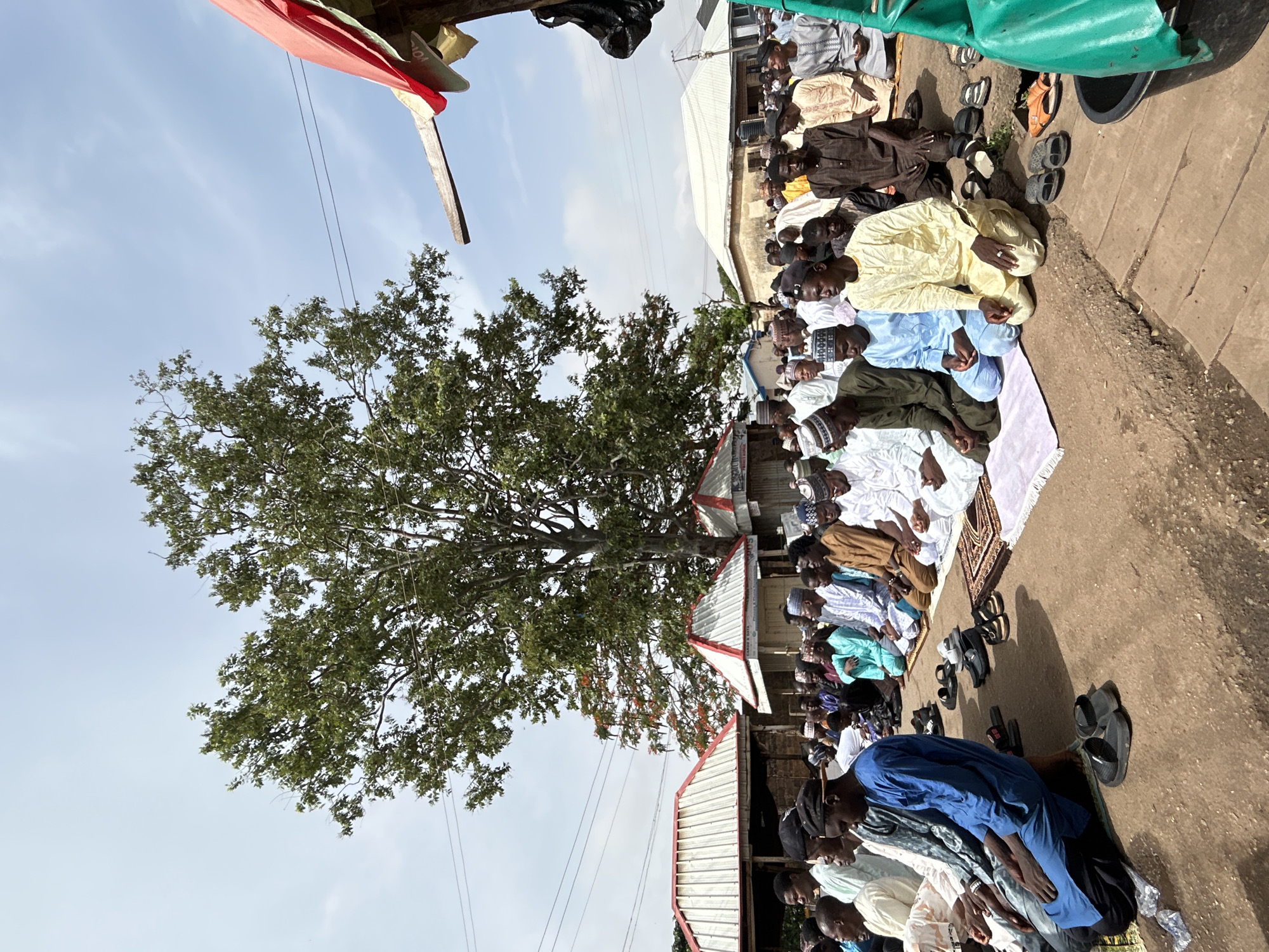 A group of men are praying outdoors in Jos, Nigeria. They are kneeling on prayer mats or the bare ground, and many have removed their shoes, which are lined up nearby. The men are dressed in a variety of traditional clothing, including robes and caps. A large tree provides shade for the worshippers. Some buildings are visible in the background.