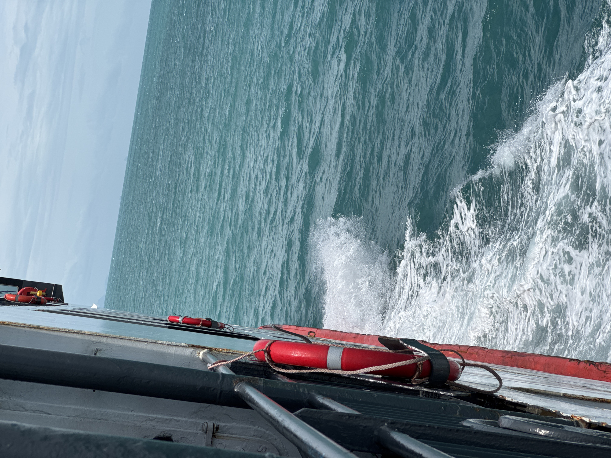 The image captures a side view of a ferry sailing through turquoise waters under a cloudy sky. The ferry's dark hull and red trim are visible along the left side of the frame. Two orange life preservers are attached to the side of the ferry, one prominently displayed with a rope. White foam and spray are generated by the ferry's movement, creating a wake in the ocean. In the distance, a faint landmass or other vessels can be observed on the horizon. The overall environment suggests a maritime setting, likely off the coast of Thailand given the context provided.