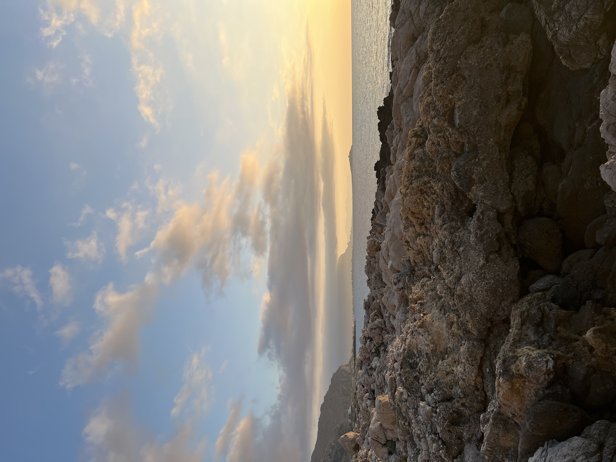 This is a vertical, eye-level shot of a rocky coastline at sunset, looking out over the sea. The foreground is dominated by large, rough, tan and grey rocks. In the middle ground, the sea stretches out to the horizon, with a faint line of land visible in the distance, possibly mountains. The sky above is a gradient of blue and yellow, with scattered, fluffy clouds tinged with orange and pink from the setting sun. The lighting suggests it is either sunrise or sunset. There are no people or animals visible in the image. The scene is serene and natural, with no man-made structures immediately apparent in the foreground or middle ground. The presence of a distant coastline and island suggests a coastal location. The city of Palermo, Italy, is known for its rugged coastline and islands, making it a plausible setting for this image.