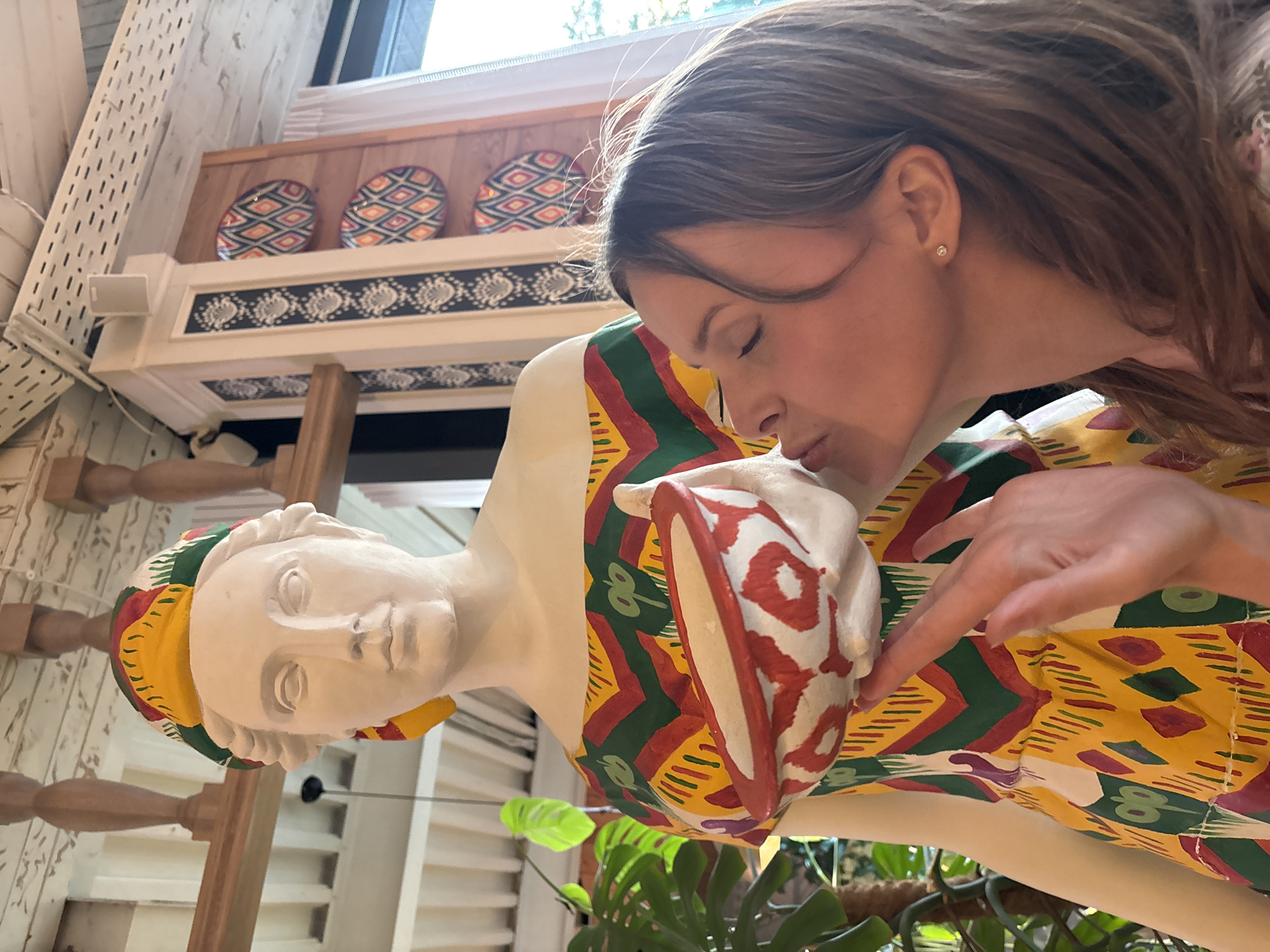 In Solnechnoye, Russia, a young woman with shoulder-length brown hair and closed eyes is leaning in to kiss a statue of a woman. The statue is pale and appears to be made of plaster, wearing a colorful dress with a geometric pattern of yellow, green, and red. The statue also sports a matching headscarf. In its hands, the statue holds a bowl decorated with red and white patterns. The background suggests an indoor environment with wooden beams and decorative plates on the wall. A large green plant is visible to the left of the statue. The lighting indicates it is daytime.