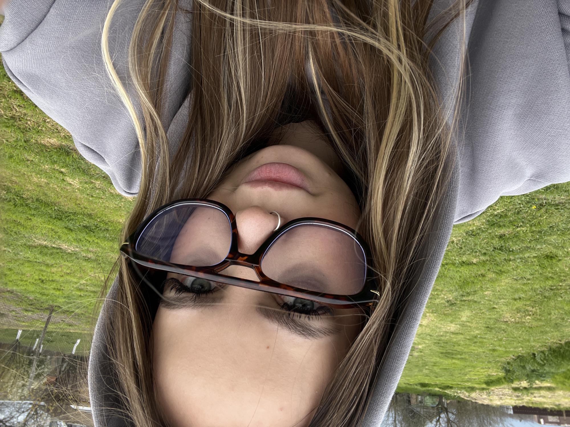 A selfie image shows a young woman wearing a grey hooded sweatshirt and tortoiseshell-framed glasses. Her hair is blonde and brown, partially obscuring her face. She has a silver nose ring. The background shows green grass and trees, suggesting a park or outdoor area. Location information is unavailable to determine if it is the Russian city of Port-Katon.
