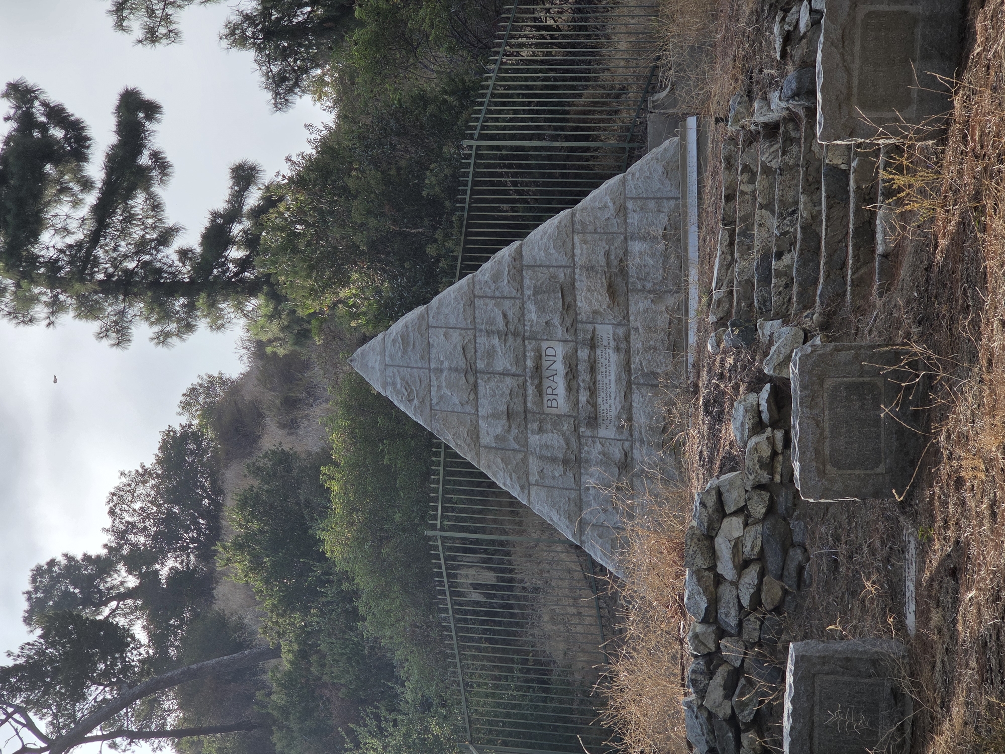 The image shows a pyramid-shaped monument with the name "BRAND" inscribed in large letters. Below this, smaller text is carved into the stone. The monument is made of light-colored stone blocks and stands on a hillside covered with dry grass and some greenery. A metal fence runs behind the monument, and a stone staircase leads up to it from the foreground. Several smaller, rectangular stone markers or headstones are visible in the foreground. The environment appears to be a cemetery or memorial site. The sky is overcast, and trees are visible on the hillside. Based on the monument and its context, this is likely the Brand Park Monument in Glendale, California, United States of America, dedicated to Leslie Brand, an early resident and founder of the city.
