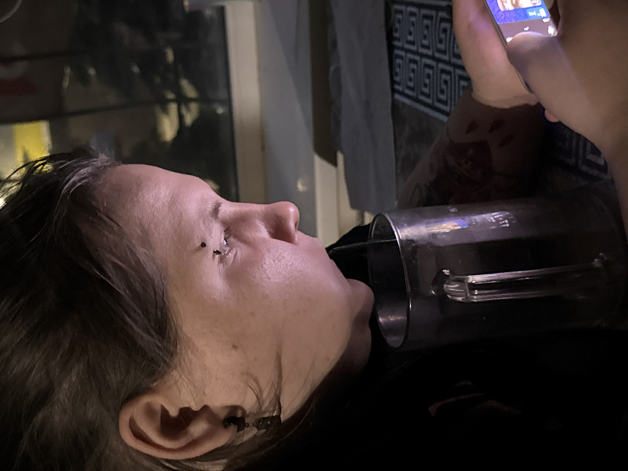 In a dimly lit indoor setting, a person is captured from the chest up, leaning forward to drink from a large, clear mug with a handle. The mug contains a dark liquid and a straw. The person's head is turned to their right, and they appear to be looking at a smartphone held in their right hand. Facial piercings are visible near the person's eye. The person is wearing dark clothing. In the background, a window offers a view of dark foliage, suggesting an outdoor environment with trees. The overall ambiance is casual and personal. The location within the city of Zaokskiy, Russia, is not discernable from the visual information provided in the image.