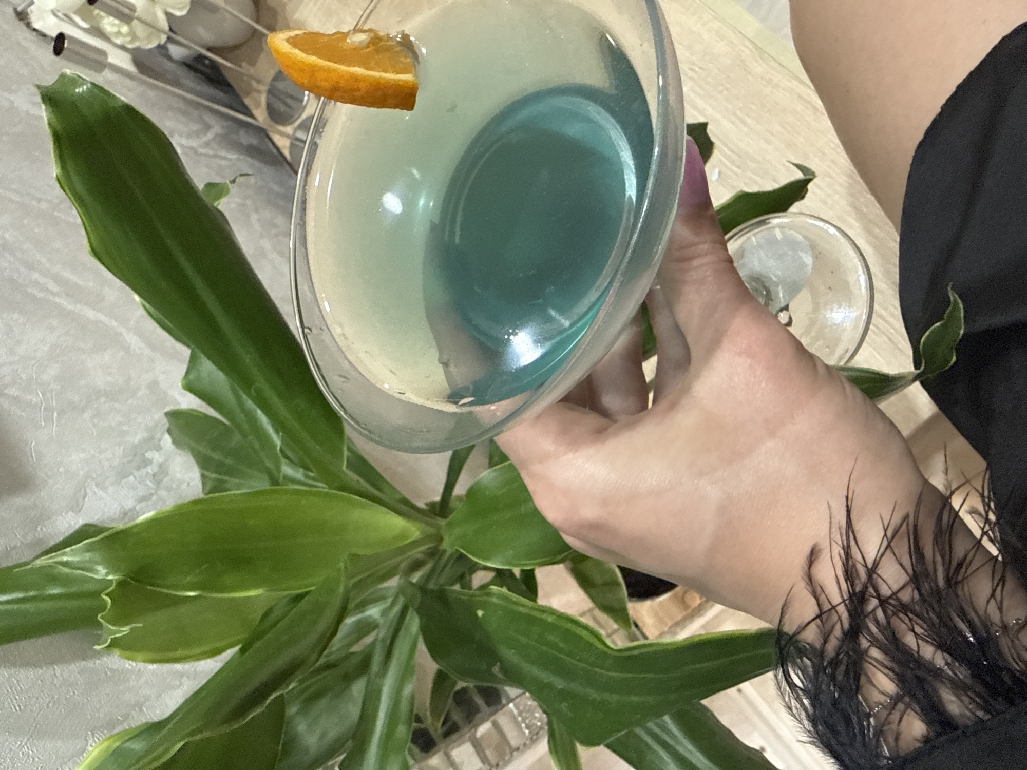 The image shows a person holding a martini glass containing a blue cocktail with an orange slice garnish. The person is wearing a black garment with feathered accents on the wrist. A green plant with long leaves is visible in the background, along with a textured wall. The scene appears to be indoors, possibly a home or restaurant setting. The location of Октябрьское, Russia, is not discernible from the visual information provided in the image.